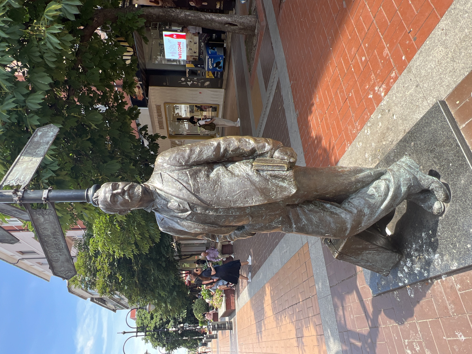 The image captures a bronze statue of a man in a suit, standing next to a suitcase and holding a book. The statue is located on a paved pedestrian street in Sofia, Bulgaria, likely in the city center. In the background, there are buildings with shops, trees lining the street, and a clear blue sky. Several people are visible walking or sitting on benches in the background, suggesting a public area. One signpost above the statue has directional arrows with text in Cyrillic script.