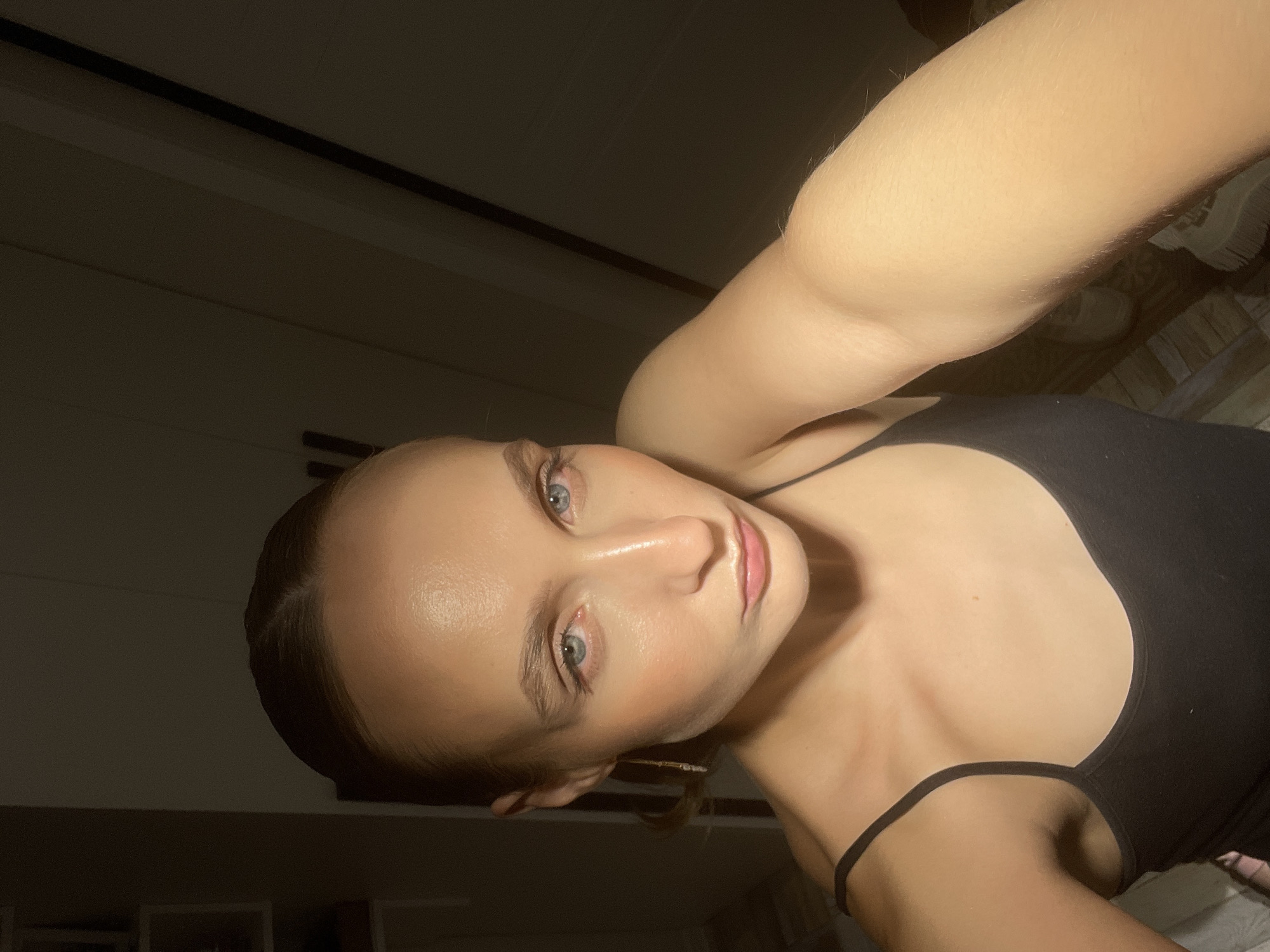 The media file is a close-up, low-angle selfie of a young Caucasian woman. She has fair skin, light blue eyes, and light brown hair pulled back. She is wearing a black tank top. The lighting is dim, suggesting it might be evening or nighttime. The background is dark and appears to be an indoor setting, possibly a bedroom, with hints of wooden paneling on the wall or furniture. The woman's arm is extended, holding the camera, and her gaze is directed towards it. Her expression is neutral or slightly thoughtful. There is no visible text in the image. The location context provided is Chaikovsky, Russia.
