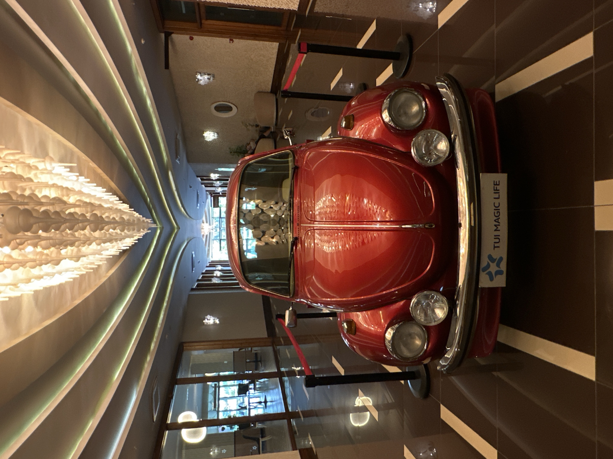 A bright red vintage Volkswagen Beetle is positioned centrally in a hotel lobby hallway, oriented front-facing. The car is roped off with red velvet stanchions and tape. The hallway features a polished, dark brown tiled floor with light-colored stripes. The ceiling has a distinctive, modern design with a large, elaborate chandelier and recessed lighting along curved beams. Large glass doors and windows line the left side of the hallway, offering glimpses into other hotel areas. The right side of the hallway has textured walls with decorative light fixtures. The environment appears to be indoors, likely a hotel or resort, and the car is a decorative display. The plaque on the car reads "TUI MAGIC LIFE". The location is identified as Manavgat, Turkey, suggesting this scene is within a TUI Magic Life resort in that region. There are no people clearly visible in the immediate vicinity of the car.