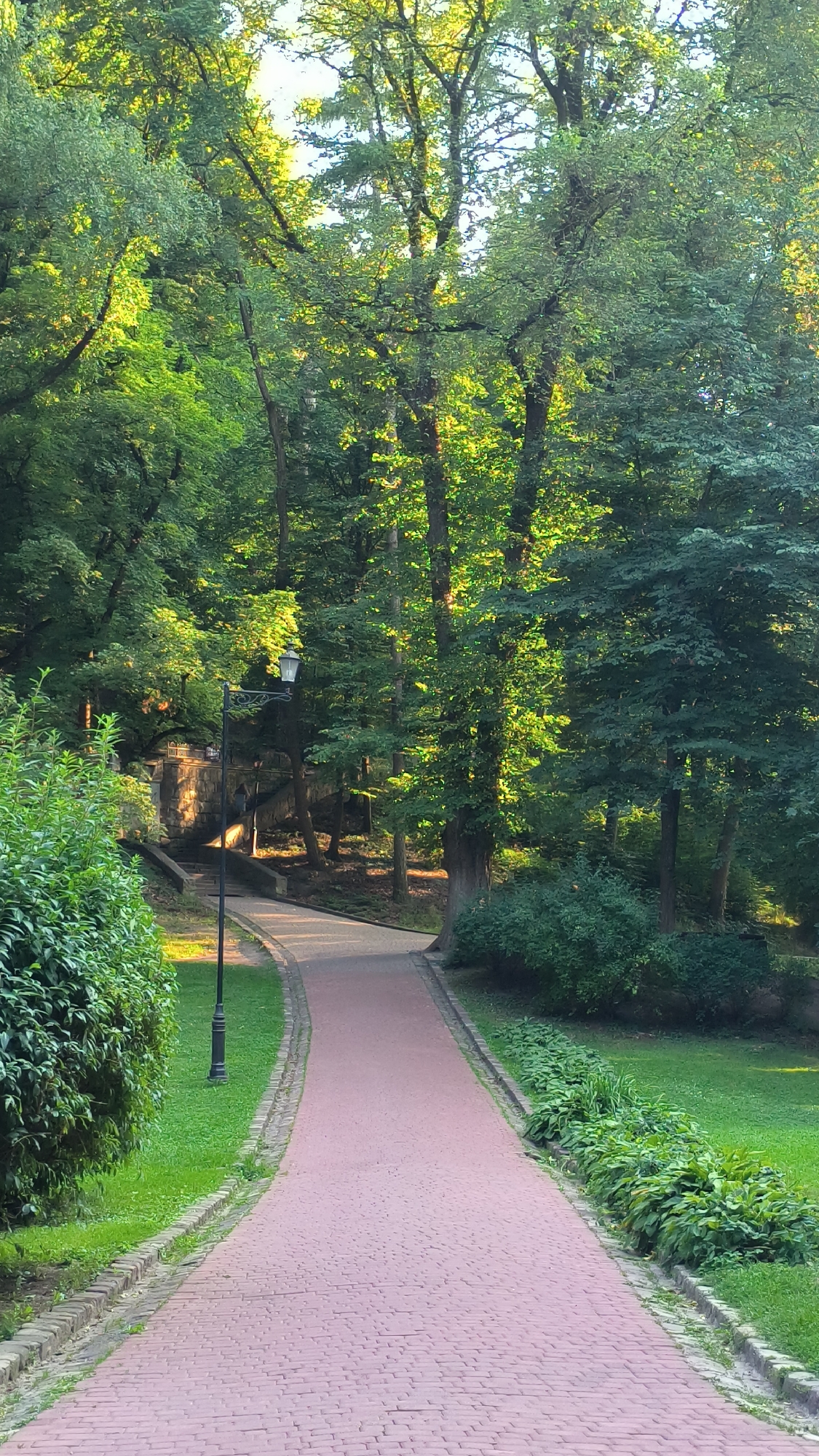 A paved pathway curves through a sun-dappled park, lined by lush green grass and trees. The pathway is made of reddish-brown bricks, bordered by a low brick edge and strips of grass. A vintage-style lamppost stands on the left side of the path, near a stone staircase that leads up a gentle incline. The staircase has a stone retaining wall on one side. Dense foliage, primarily trees with green leaves, forms a canopy overhead, allowing shafts of sunlight to illuminate parts of the scene. In the distance, on the left side of the frame, a stone structure is visible beyond the staircase, partially obscured by trees and shrubs. The overall scene depicts a tranquil park setting, likely in Lviv, Ukraine, possibly in the area of Stryiskyi Park given its characteristic pathways, lampposts, and mature trees.