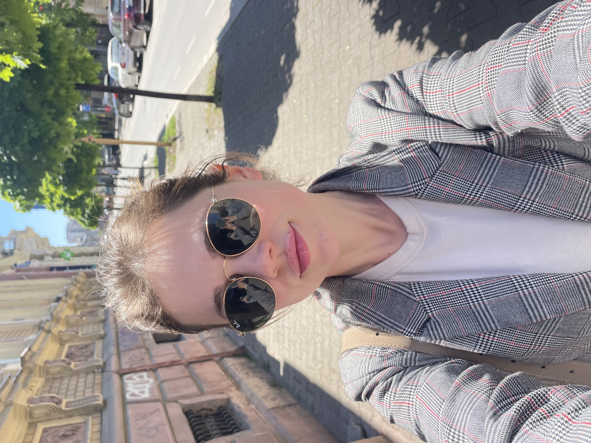 A selfie depicts a person wearing sunglasses and a plaid blazer on a city street. The person is positioned at an angle, facing towards the camera. Behind the person, there is a building with detailed architectural features and a street with cars parked alongside. The scene is set in Novi Sad, Serbia, based on the architecture and general cityscape.