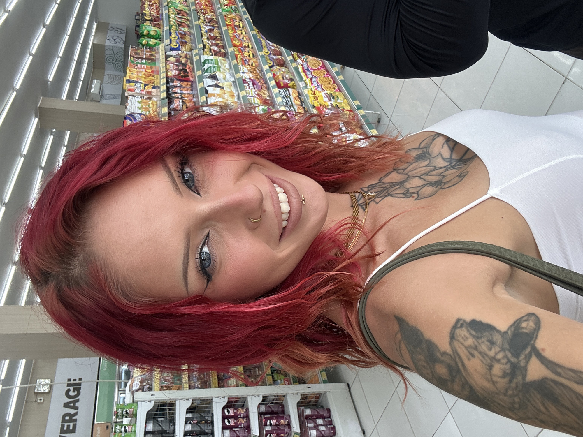A red-haired woman with tattoos on her neck and right arm smiles into the camera. She wears a white top and is in a store with shelves of items visible in the background. A person wearing black clothing is partially visible above her. "Beverage" is partially visible on a shelf in the background. The location is likely a grocery store, and the city is Kuta Selatan, Indonesia.
