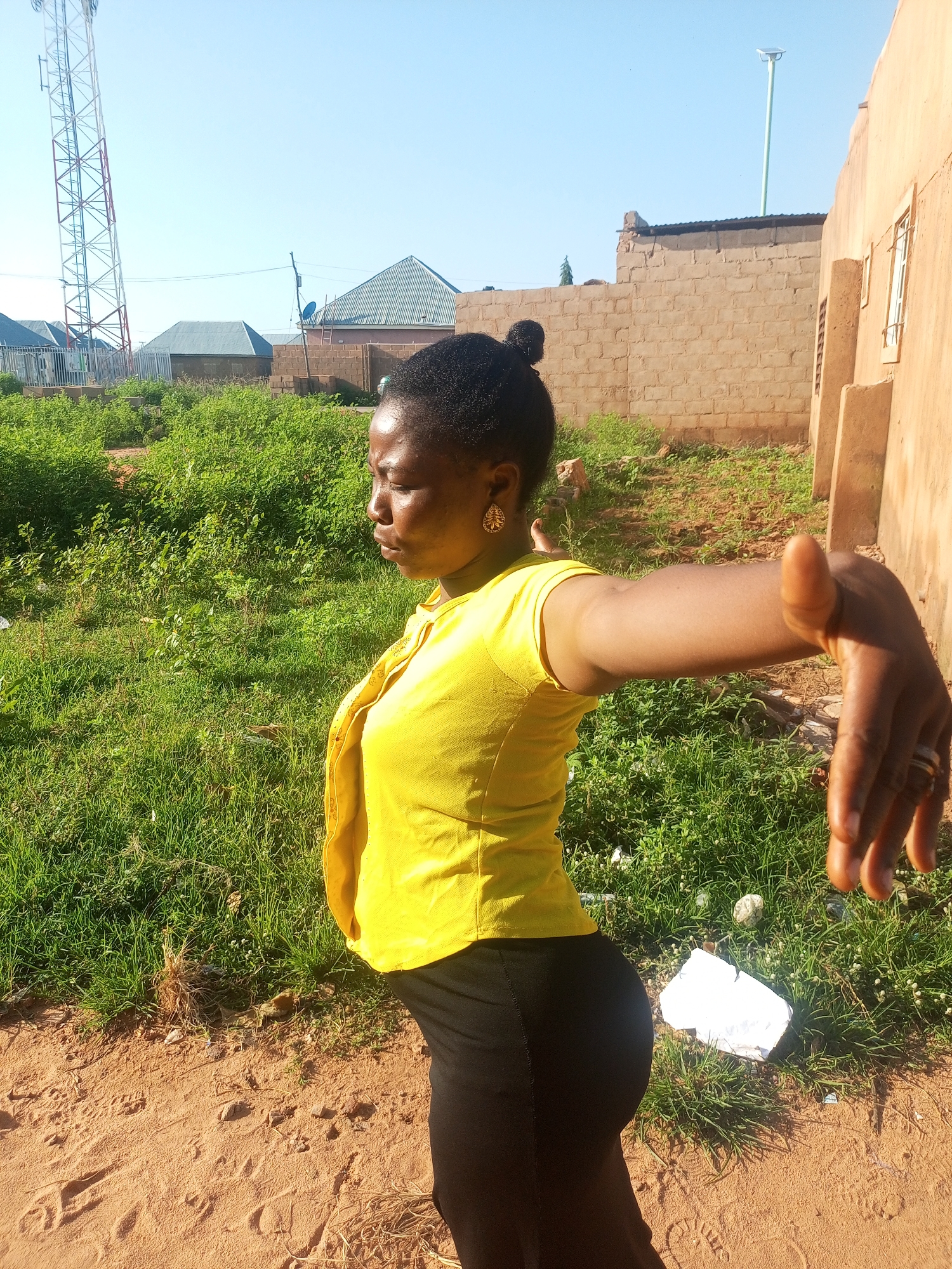 A medium shot captures a Black woman standing in front of a brick building, her body angled in profile to the left. She has her right arm extended forward, with her hand in a fist and thumb pointed up. She is wearing a bright yellow short-sleeved shirt and black pants. Her hair is pulled back in a bun. The background shows a clear blue sky, a green grassy area with some scattered trash, and a cell tower in the distance. The ground in the foreground is sandy and reddish-brown. The scene appears to be outdoors in a semi-urban or rural area.