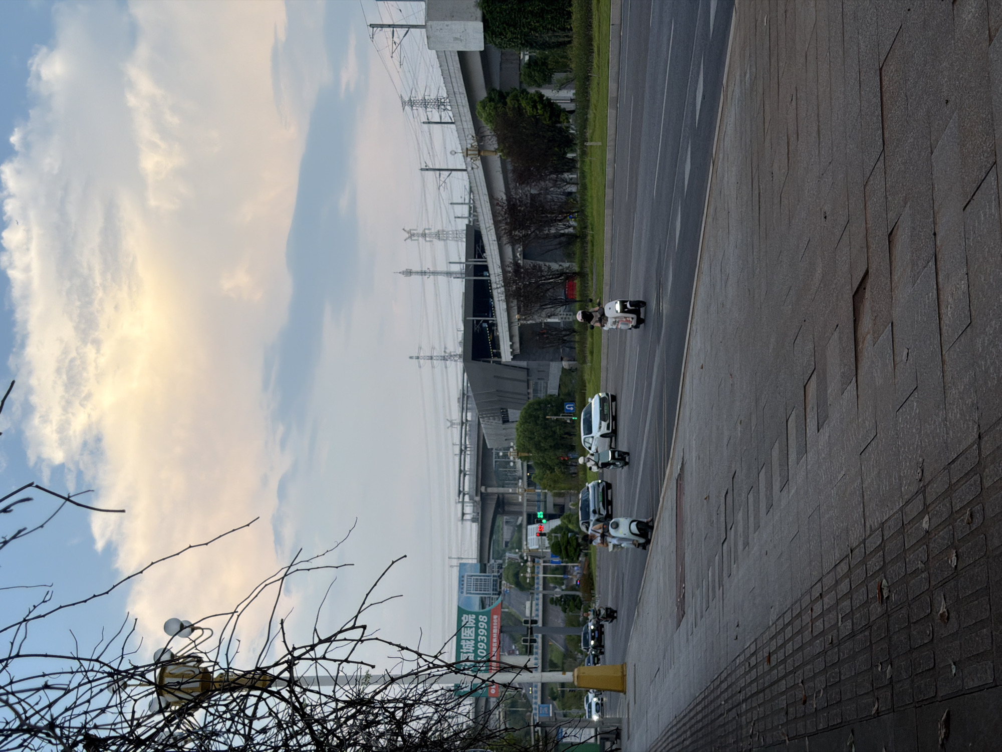 This is an outdoor, eye-level, medium shot photograph of a street in Jiangjin, China. The image captures a road with multiple lanes, a sidewalk, and an elevated structure that appears to be a highway or train line in the background. The sky is partially cloudy with some soft, yellowish light suggesting either sunrise or sunset. A street lamp is visible on the left side, partially obscured by tree branches. Several vehicles, including cars and scooters, are on the road. On the sidewalk, there are textured paving stones and scattered leaves. In the distance, there are power lines and tall transmission towers. A billboard with Chinese text and a phone number is also visible. The overall impression is of an urban environment during twilight.