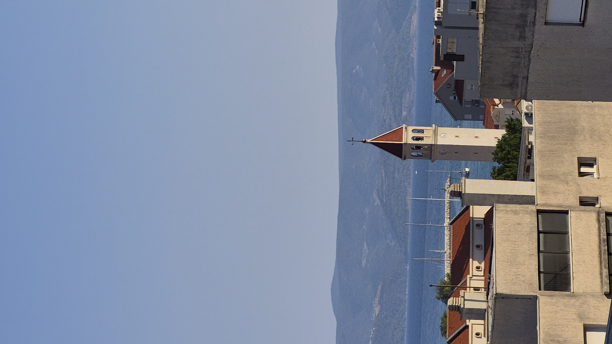 A high-angle, outdoor, eye-level shot captures a distant view of a coastal town. The sky is clear and blue, transitioning to a hazy blue horizon over a body of water. A mountain range forms the distant backdrop, its contours softened by atmospheric conditions. In the middle ground, the blue water is dotted with sailboats, their masts reaching skyward. The foreground is dominated by the rooftops and upper stories of buildings. A prominent church bell tower, with a red-tiled, conical roof and a cross at its apex, stands out among the other structures. The bell tower features arched openings and a clock face. Adjacent buildings have red-tiled roofs and tan-colored facades. The overall scene depicts a bright, sunny day in a Mediterranean-style coastal settlement.