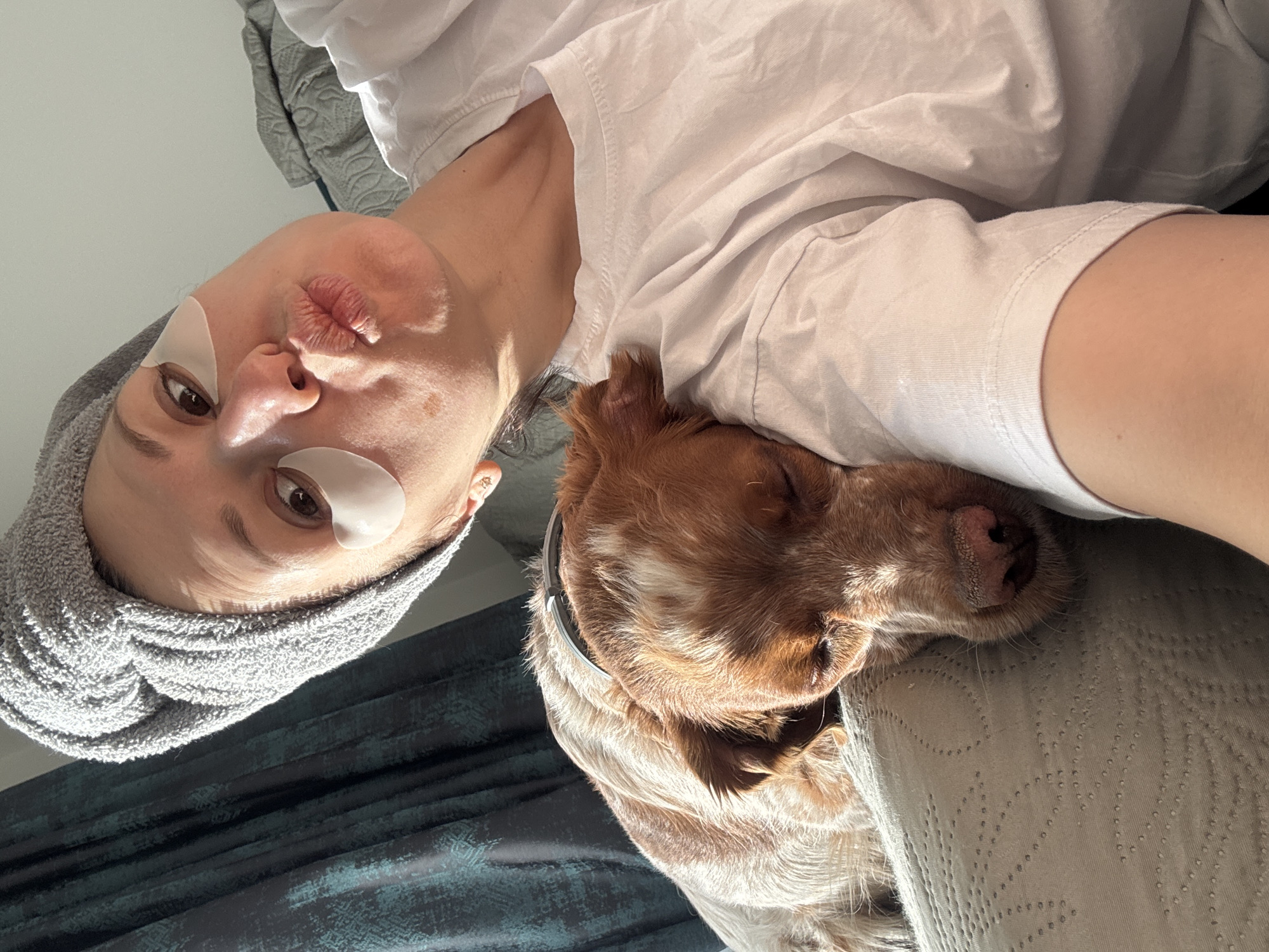 A woman is taking a selfie indoors. She has a gray towel wrapped around her head and white eye patches under each eye. She is making a kissing face at the camera. She is wearing a white t-shirt and resting her head on a pillow. A brown and white dog with long ears is resting its head on the woman's shoulder. It is not possible to determine the location is Sofia, Bulgaria based on the image.