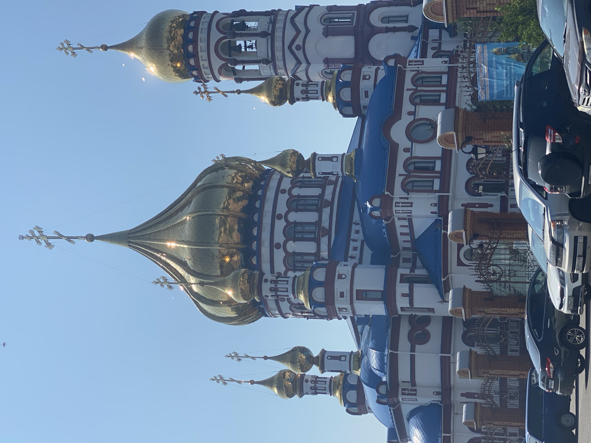 The image shows the Novokuznetsk Cathedral of the Nativity of Christ, a prominent landmark in the city of Novokuznetsk, Russia. The cathedral is characterized by its multiple golden domes, intricate white and blue architecture, and red trim. The sky is clear and blue. In the foreground, several cars are parked, and a decorative fence surrounds the cathedral grounds. There are no people visible in the image. The scene is bathed in sunlight, suggesting a clear day.