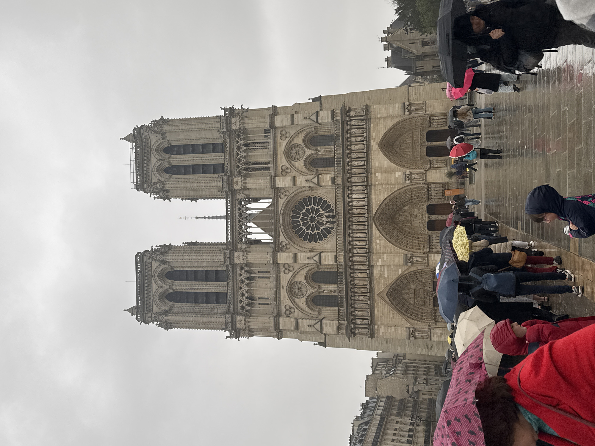The image captures the facade of Notre Dame Cathedral in Paris on a rainy day. The architectural details of the cathedral, including its towers, rose window, and arched doorways, are visible. A group of people holding umbrellas stand in front of the cathedral, possibly waiting in line. The ground is wet, reflecting the overcast sky. Other buildings are visible in the background, indicative of the urban environment.
