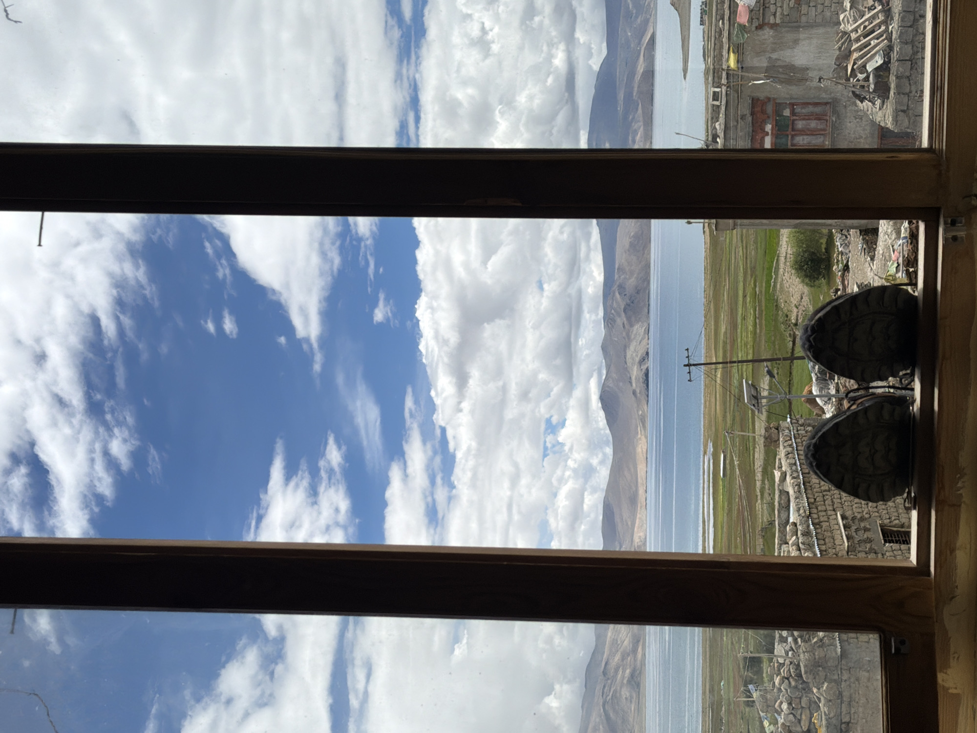 The image shows a view through a wooden window frame, with two dark-colored shoes resting on the windowsill in the foreground. Beyond the window, a large, calm body of water stretches towards a mountainous horizon under a bright blue sky dotted with fluffy white clouds. The middle ground features a green, grassy landscape with some scattered buildings and structures, possibly a village. The right side of the image reveals more of the village with stone buildings, some with red-trimmed windows, and debris piled up. This view is consistent with the location in Karzok, India, a village situated on the shores of Tso Moriri lake in Ladakh.