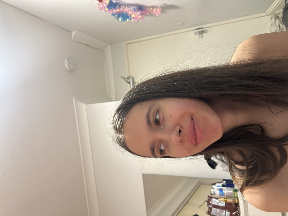The image displays a young female subject indoors, located in Corby, United Kingdom. The subject, positioned slightly to the right of the frame's center, possesses long, straight, dark brown hair and is looking towards the upper left with a subtle smile. Her right shoulder is bare. The room features white walls and ceiling with white crown molding. A white smoke detector is affixed to the ceiling in the upper right quadrant. A light-colored door with a silver handle is visible to the right. A decorative pink and blue tinsel unicorn figure is mounted on the wall to the right of the door. The left background shows a partial mirror reflection of various bottles and a brown package on a surface. Some undefined clutter is visible on the floor in the lower right.