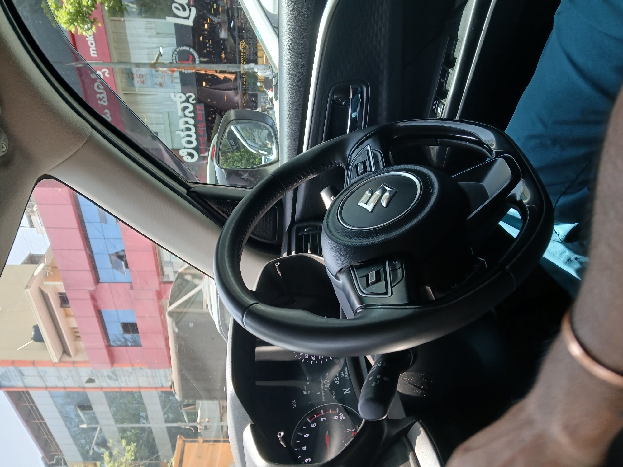 The image shows the interior of a car, specifically the driver's side, with a Suzuki steering wheel prominently featured. Through the windshield, a street scene is visible. Signboards in Kannada script, alongside signs like "Le Wings Burger" and "Make," indicate the image was taken in Bengaluru, India. A man, presumably the driver, is seen in a blue shirt with a metallic bracelet on his wrist. The car's dashboard displays information such as average fuel economy (14.2 km/L) and range (43 km). The odometer reads "34928km." The gearshift is in neutral. The surrounding buildings are visible through the windshield.