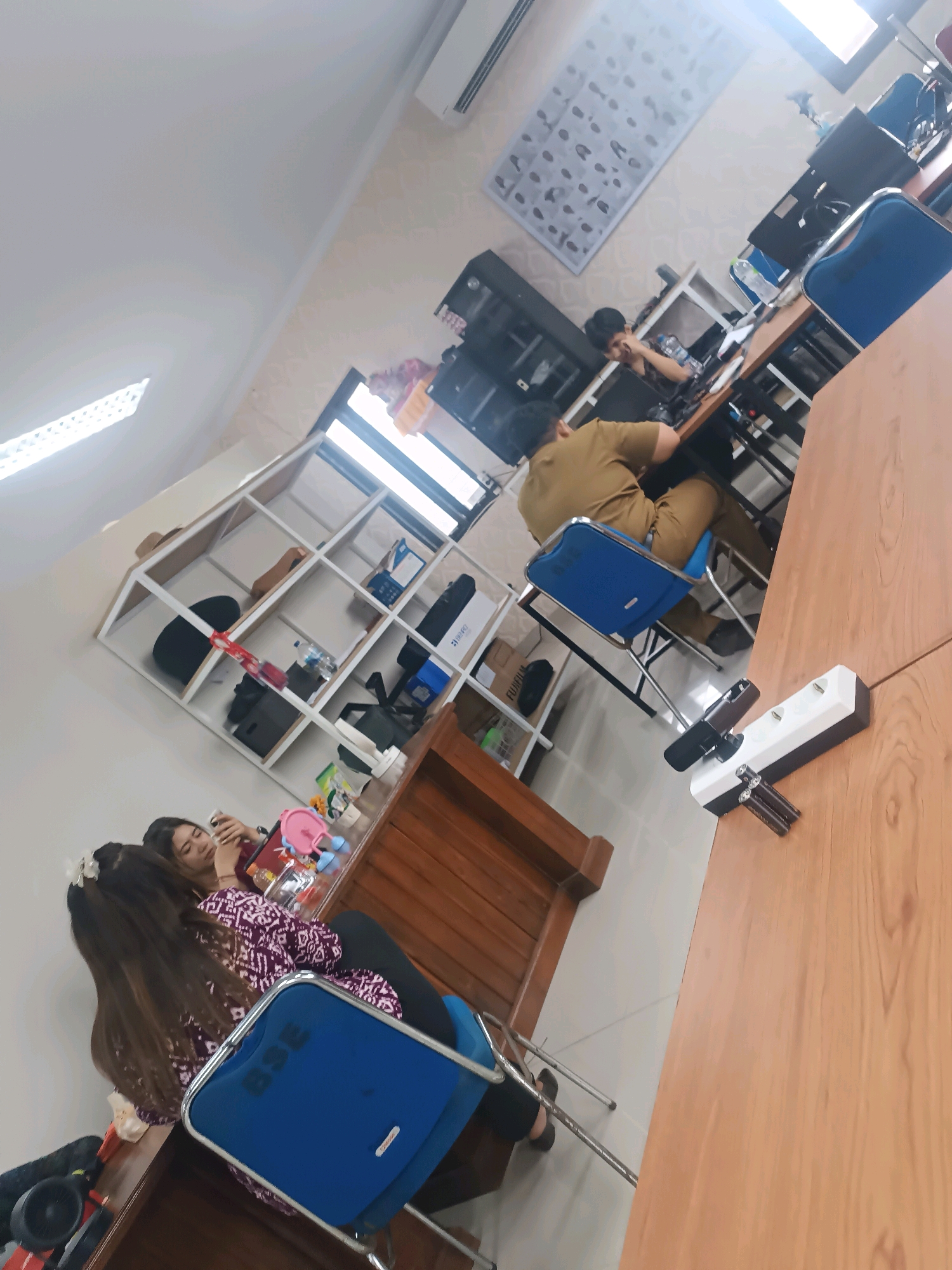 The image depicts an indoor scene, possibly an office or a classroom, with several people and objects. The room has white walls and a light-colored floor. In the background, there are shelves filled with various items, including boxes, bottles, and bags. A man in a brown uniform is seated at a desk in the middle ground, appearing to be working on a computer. To the left, two women are seated at a table, with one holding a phone. A blue chair is visible in the foreground, as is a wooden table with a power strip and electrical outlets. The overall lighting suggests it is daytime, with natural light coming from windows. The location is specified as Tabanan, Indonesia.