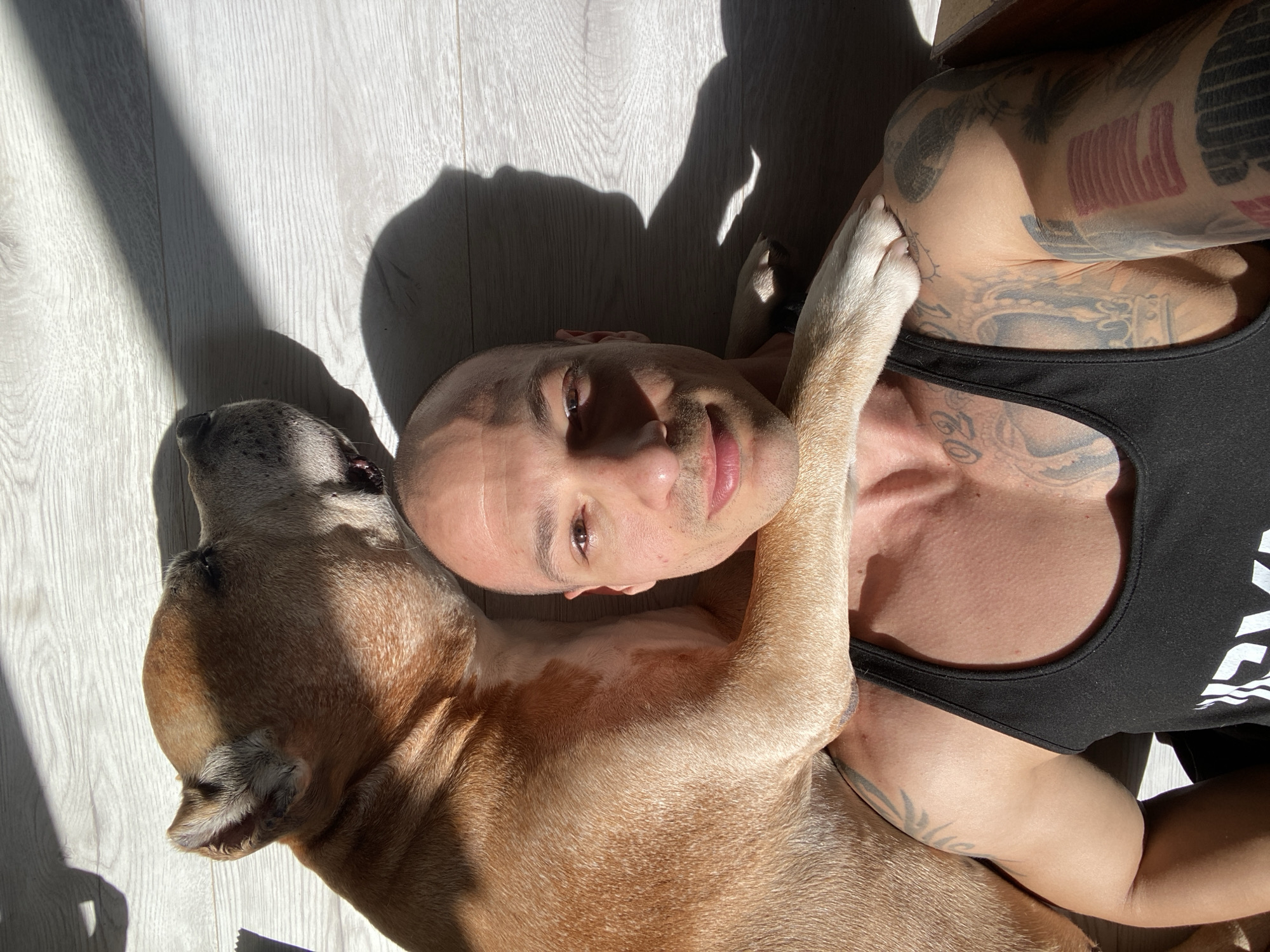 A bald man with tan skin and a black tank top lies on a light-colored wooden floor with his dog. The dog is light brown with a grey muzzle and is resting with its paw on the man's shoulder. The man has multiple tattoos on his right arm and chest. Sunlight casts a shadow across the floor. There is no specific evidence indicating this scene is located in Pleven, Bulgaria.