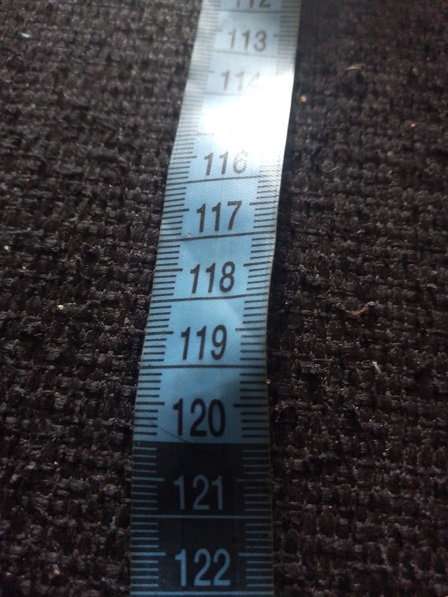 A close-up, low-angle shot shows a blue measuring tape laid diagonally across a dark, textured surface, possibly carpet or fabric. The measuring tape is marked with white numerical graduations and thin black lines. The visible numbers on the tape are 112, 113, 114, 116, 117, 118, 119, 120, 121, and 122. The surface beneath the tape is dark brown or black and appears to be a rough textile with a visible weave. The lighting is uneven, with a bright highlight on the measuring tape and shadows on the textured surface. There are no people or discernible city features present in the image. The scene does not provide any specific information to locate it in Jauro Yinu, Nigeria.