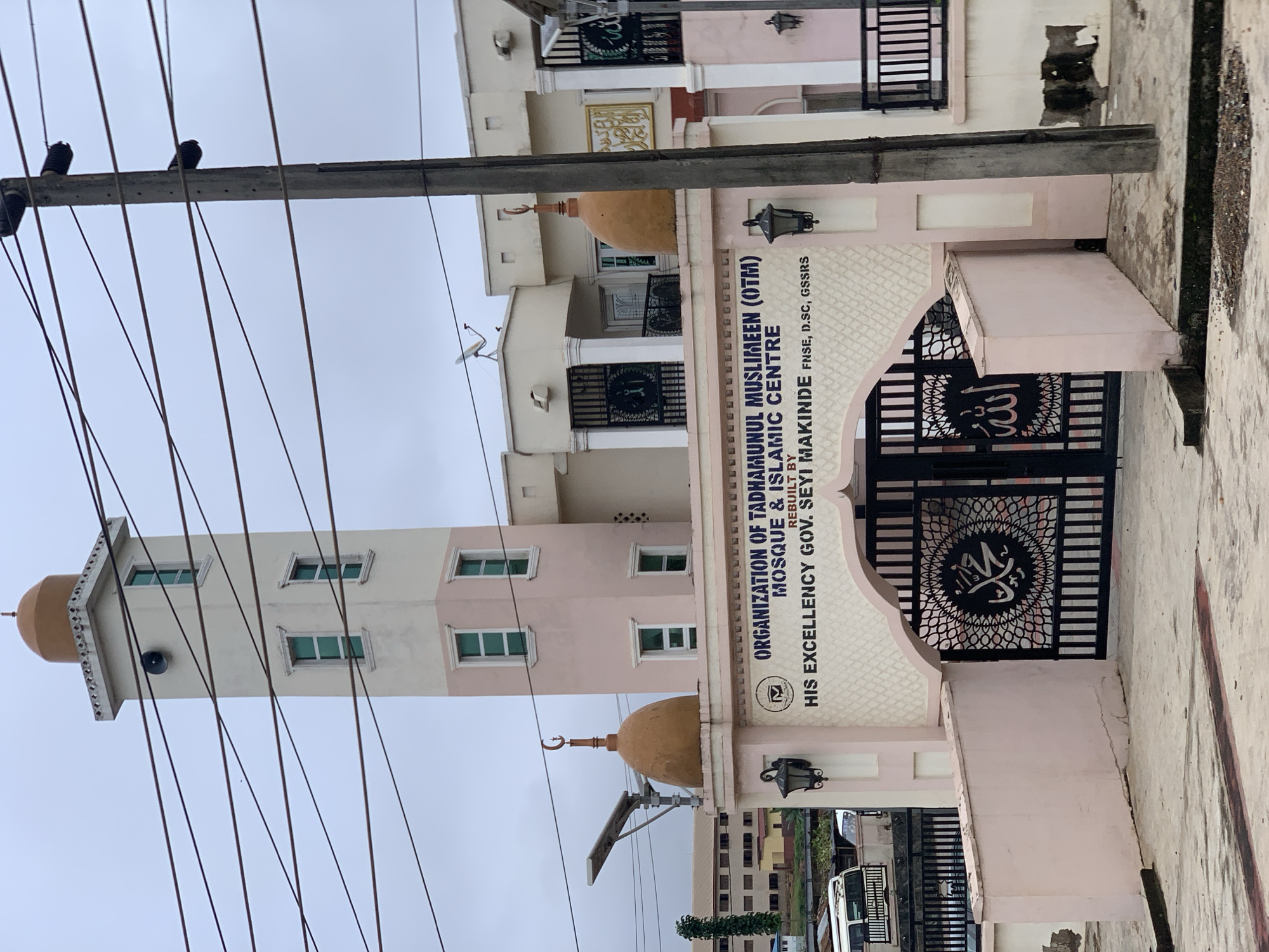 The image shows the exterior of the Organization of Tadhmanul Muslimeen (OTM) Mosque & Islamic Centre, located in Alakia, Nigeria. The building features a tall, multi-story minaret with a dome at the top, painted in light pink and white. There are several windows on the minaret and other parts of the building, some of which appear to be stained green. Two smaller domes, each topped with a crescent moon, are visible on either side of the main entrance arch. The entrance gate is made of black metal with intricate Islamic calligraphy and patterns. Above the gate, a sign reads "ORGANIZATION OF TADHMANUL MUSLIMEEN (OTM) MOSQUE & ISLAMIC CENTRE REBUILT BY HIS EXCELLENCY GOV. SEYI MAKINDE, FNSE, D.SC, GSSRS". The building is painted in pastel colors, primarily light pink and cream. A concrete utility pole with multiple electrical wires runs vertically on the right side of the image. The ground in front of the mosque is paved concrete with some areas of dirt and debris. In the background, other buildings and structures are visible, along with a cloudy sky.