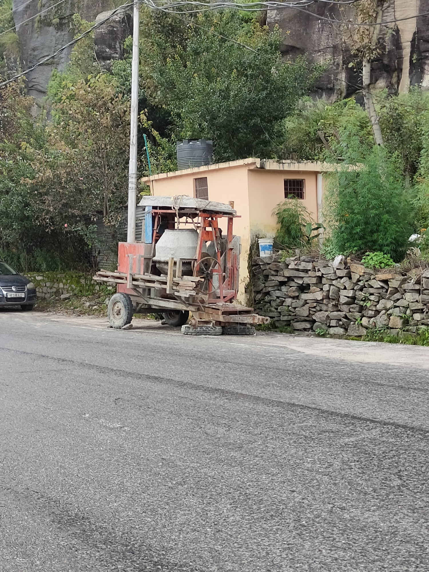 The image captures a construction site scene in Barheta, India. In the foreground, a textured asphalt road is visible. To the right, a dry stone wall forms a boundary, topped with green vegetation. Behind the wall, a small, peach-colored building with a barred window stands. A dark gray car with a license plate reading "HP 588 6051" is parked on the left side of the road. The central focus of the image is a red and silver portable cement mixer, equipped with a large drum and a metal wheel for manual operation. The mixer is resting on a trailer with two wheels, one of which is visible. Several wooden planks are stacked on the trailer. In the background, lush green trees and a rocky cliff face dominate the landscape. A tall, slender utility pole stands prominently near the mixer. A dark, large water tank is situated on the roof of the peach-colored building.