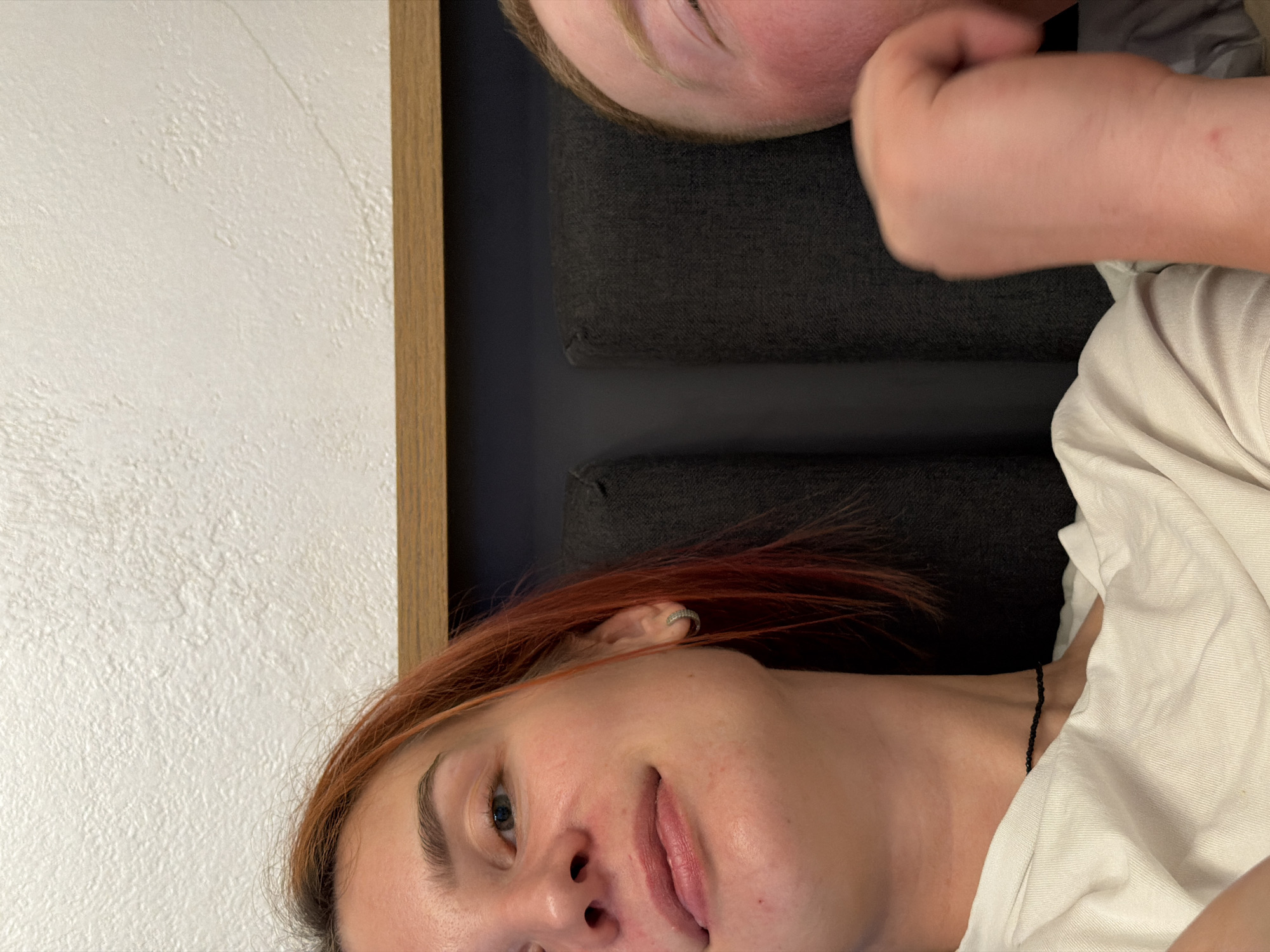 Two individuals are lying down next to each other. One person has red hair and is wearing a white shirt. The other person's face is partially visible. The location of the scene is unknown.
