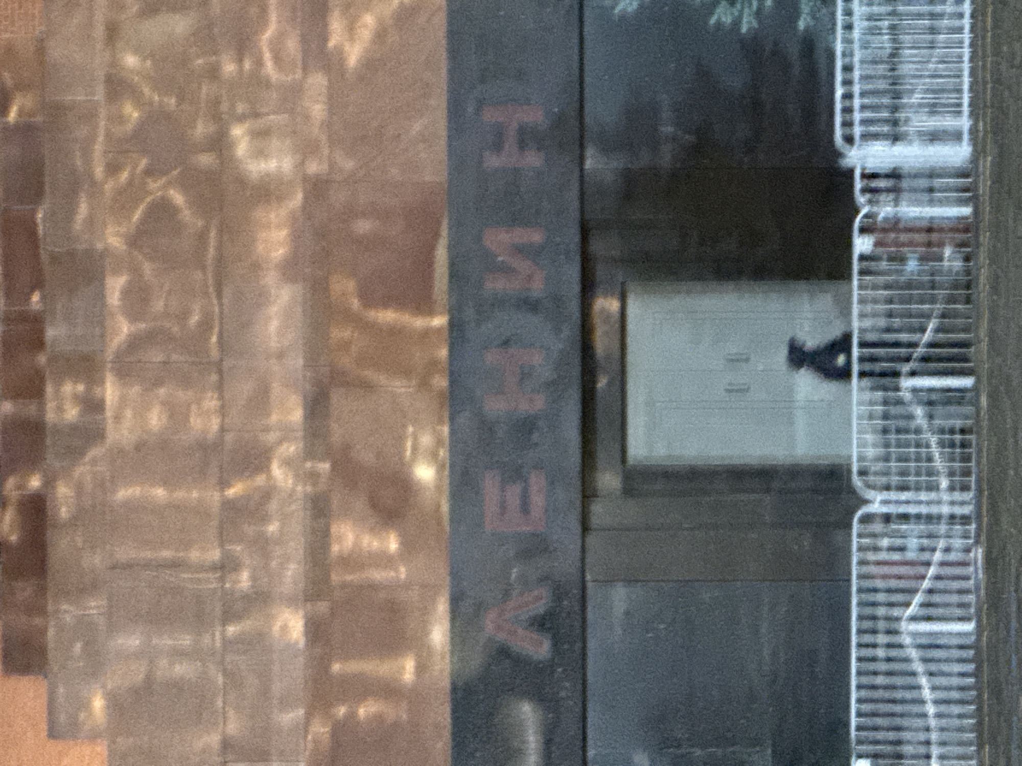 The image depicts a section of the Lenin Mausoleum in Moscow, Russia. The words "ЛЕНИН" (Lenin) are vertically inscribed in Cyrillic on a dark stone surface. Adjacent to the stone is a wall of what appears to be polished stone tiles. A reflection on the tiles shows light patterns. A blurred figure of a person appears to be walking behind a partially visible railing near a grey wall with a closed door.
