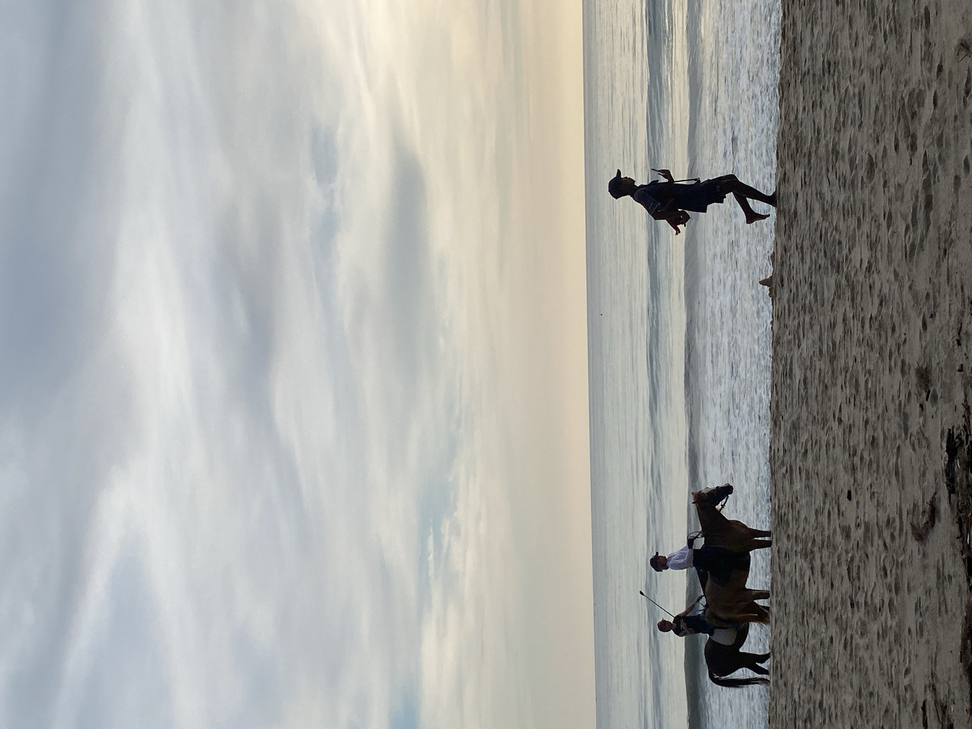 The image depicts a beach scene in Choeng Thale, Thailand. The sky is cloudy, and the water meets the sand. Two people are riding horses along the shoreline. Another person is running barefoot on the sand near the water.