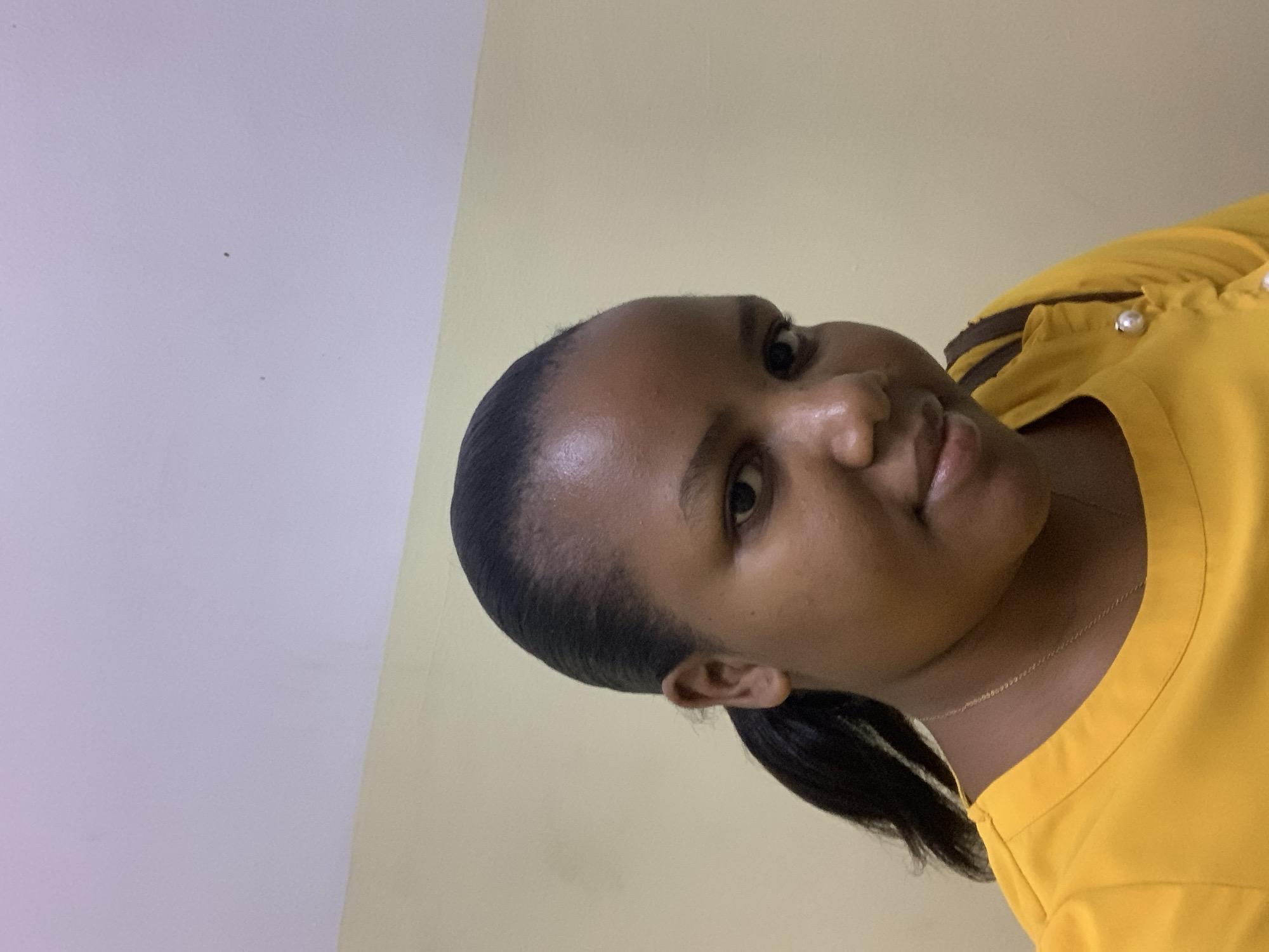 A woman is pictured in a close-up, slightly tilted portrait. She has dark hair pulled back into a low ponytail. She is wearing a yellow shirt with pearl-like buttons. A thin gold necklace is visible around her neck. The background consists of two wall colors, a light beige or yellow and a light gray or white. The photo appears to have been taken indoors. There is no evidence to suggest this photo was taken in Dar es-Salaam, Tanzania.
