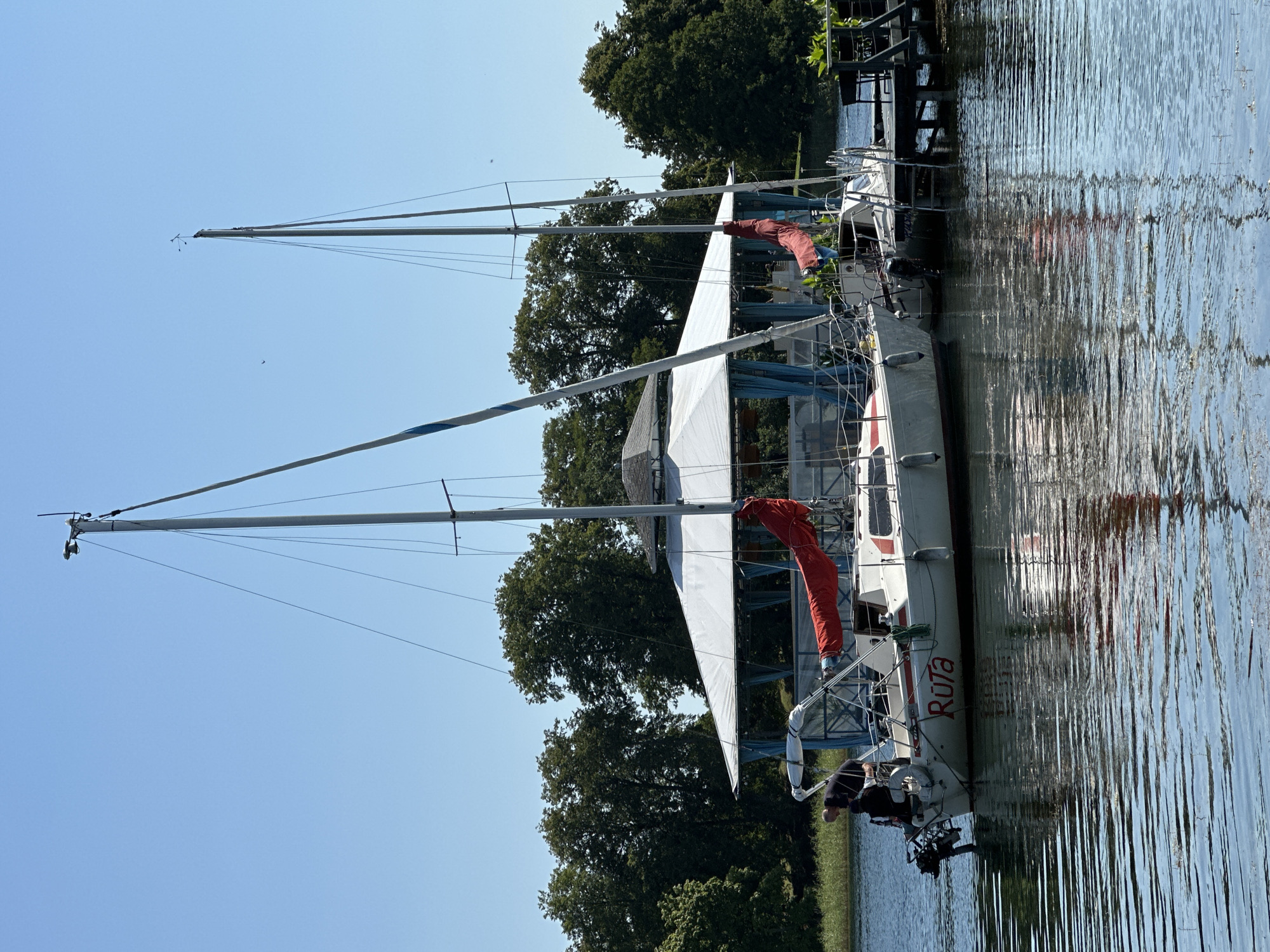 A daytime, eye-level medium shot captures two white sailboats moored on a calm body of water near a covered pier structure. The boat on the left is closer to the viewer, its hull displaying the red name "Rūta". Two individuals are visible on the deck of the "Rūta" sailboat; one person wearing a dark shirt is attending to equipment at the stern, and another person, also in dark attire, is seated. A third individual is seen on the deck of the second sailboat, which is docked to the right of the first. The pier, with a light-colored, tent-like roof and blue curtains, is situated behind the sailboats. The background consists of dense green trees on the shore, with a clear blue sky above. The scene is set in Trakai, Lithuania, likely along the shore of Lake Galvė.