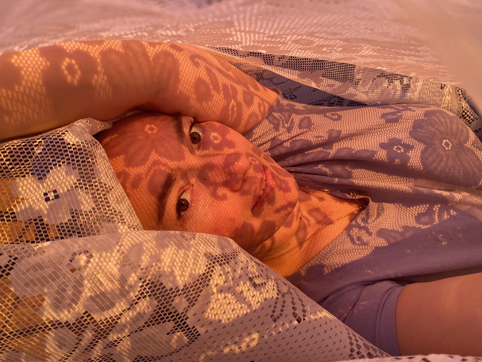 This close-up portrait features a young person, likely female, lying down in an indoor setting, possibly on a bed or couch, in Ольховка, Russia. The scene is bathed in warm, golden light, characteristic of either sunrise or sunset, creating long, intricate shadow patterns.
A white, patterned lace fabric is draped over the person and partially covers the frame. This lace casts delicate, dappled shadows across her skin, including her face, arm, and neck, as well as onto her light purple top. Another patterned fabric, also purple with a subtle floral or geometric design, is visible underneath the lace, forming part of her clothing or bedding. A thin golden chain is visible around her neck.
The person looks directly towards the viewer with open, calm eyes and a soft, gentle smile, conveying a sense of serenity and relaxation. The interplay of light, shadow, and varied fabric textures is a prominent visual element. No visible text is present in the image.
