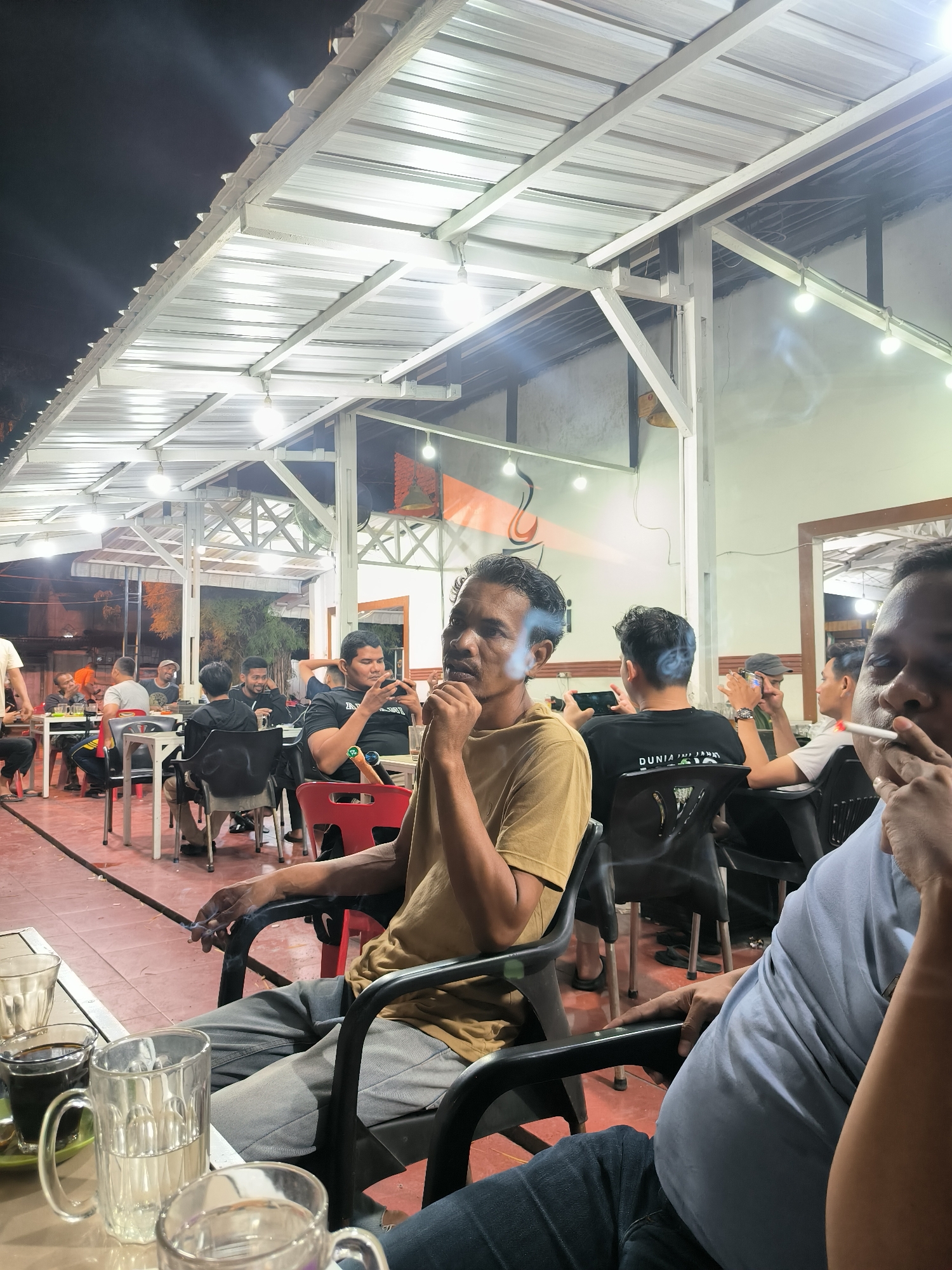 The image depicts an evening scene at an outdoor or semi-outdoor cafe in Lhokseumawe, Indonesia, illuminated by numerous overhead lights. The dark sky is visible above a prominent white metal corrugated roof structure supported by white beams.
In the immediate foreground, two men are seated, both smoking cigarettes, with plumes of smoke visible around their faces. The man on the left wears a mustard-colored shirt, while the man on the right, partially visible, wears a light blue shirt. On the table in front of them are glasses and mugs, some containing dark liquid (likely coffee) and clear liquid.
Further back, several other men are gathered at various tables with black and red chairs. They are engaged in different activities: some are talking, others are looking at their mobile phones, and several are also smoking. One man in a black T-shirt, seen from behind, has text on his shirt that reads "DUNIA INI JA..." (likely part of an Indonesian phrase). The atmosphere appears casual and social.
The floor is a reddish-brown tiled surface. In the background, beyond the main seating area, more people can be seen, and a light-colored building or wall with an orange design is visible, along with some distant trees under the night sky. The scene captures a typical evening gathering for men to relax and socialize.