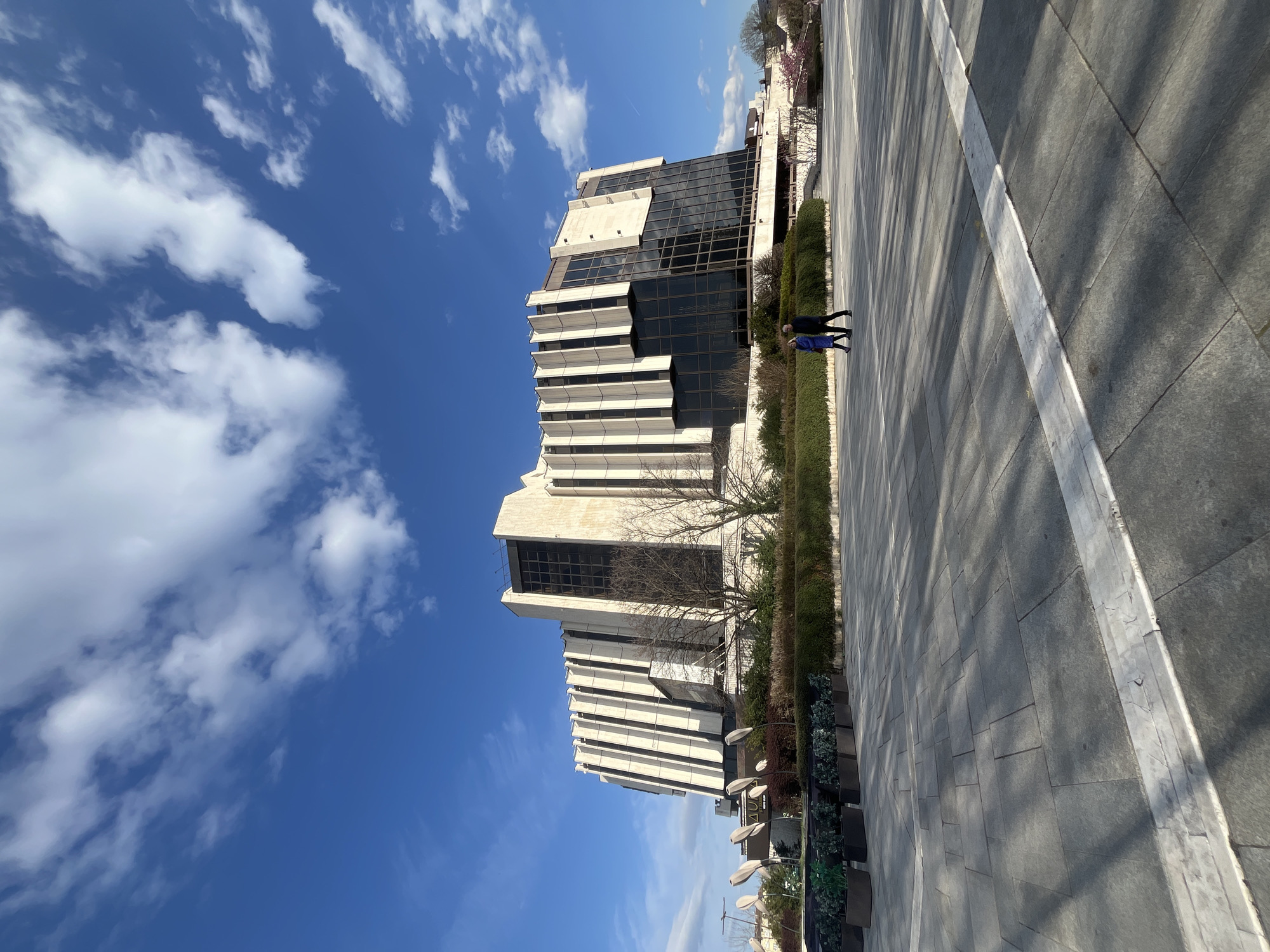 The image captures the National Palace of Culture in Sofia, Bulgaria, viewed from a ground-level perspective. The modern architectural structure with its vertical lines and dark window panes is contrasted by a vibrant blue sky dotted with fluffy white clouds.
Two pedestrians, one wearing a dark jacket and the other in a blue jacket, walk along a grey tiled public plaza adjacent to a manicured green hedge and flower boxes. The plaza is marked by a distinct white line. Bare tree branches are visible near the building. There are several modern white lampposts visible, as well.