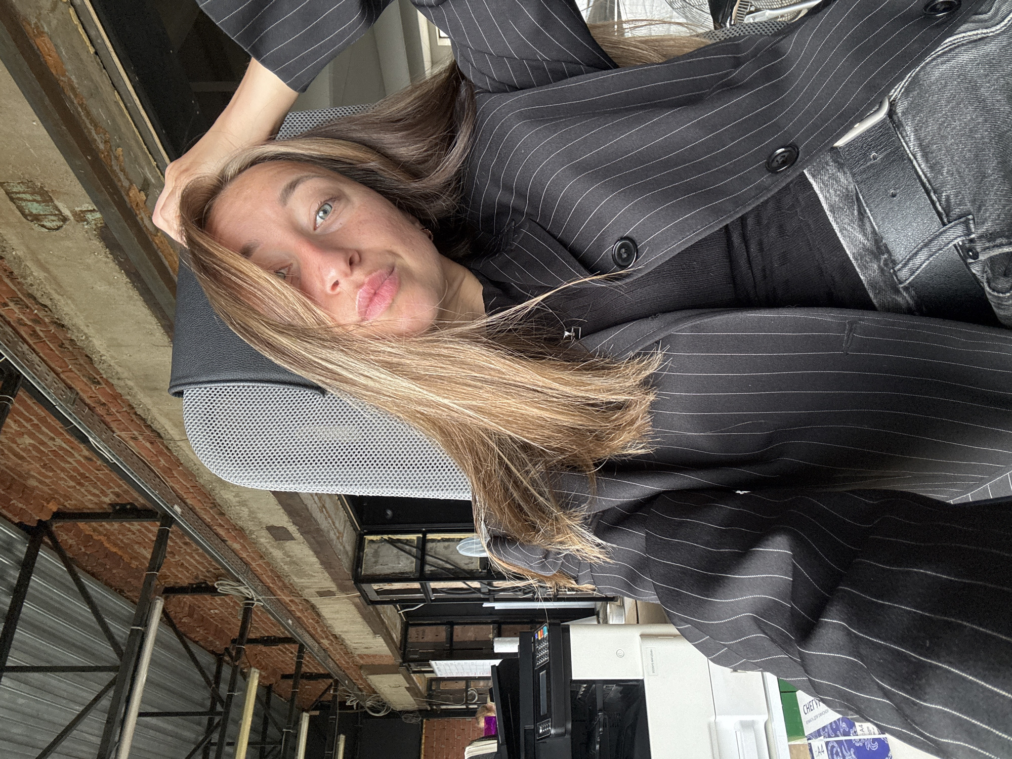 A young woman is the central subject, seen from the chest up, looking directly at the camera with blue-green eyes and slightly pouting lips. She has long, light brown hair with blonde highlights, which falls over her shoulders and part of her face. She appears to be resting her head on her left hand, which is visible near the top left of the frame.
She is dressed in a dark, possibly black, pinstripe blazer with thin white stripes, worn over a plain black top. Below the blazer, she wears dark grey or black jeans with a black leather belt and a silver buckle. Her head rests on what appears to be a grey mesh office chair.
The background suggests an indoor, possibly industrial or converted commercial space. Exposed reddish-brown brick walls are visible, along with a ceiling featuring dark metal beams, pipes, and scaffolding-like structures. Corrugated metal panels are also present in parts of the background. Various office equipment, including a white printer or copier, cables, and other electronics, are visible in the lower right portion of the image. The image is oriented diagonally, making the woman appear horizontal within the frame.
The woman is posing for a photograph, likely a selfie, given her direct gaze and relaxed yet posed posture. Her expression is calm and neutral. The lighting suggests daytime. Visible text on paper packaging includes Cyrillic script: "ЧЕРН" (possibly "ЧЕРНИЛА" - ink) and "БУМАГА ДЛЯ ПРИНТ" (BUMAGA DLYA PRINT - printer paper), along with "A4". A label on the white machine reads "F520D9-ANUAL" and "Samsung". These details, particularly the Cyrillic text, align with the location context of Chaikovsky, Russia.
