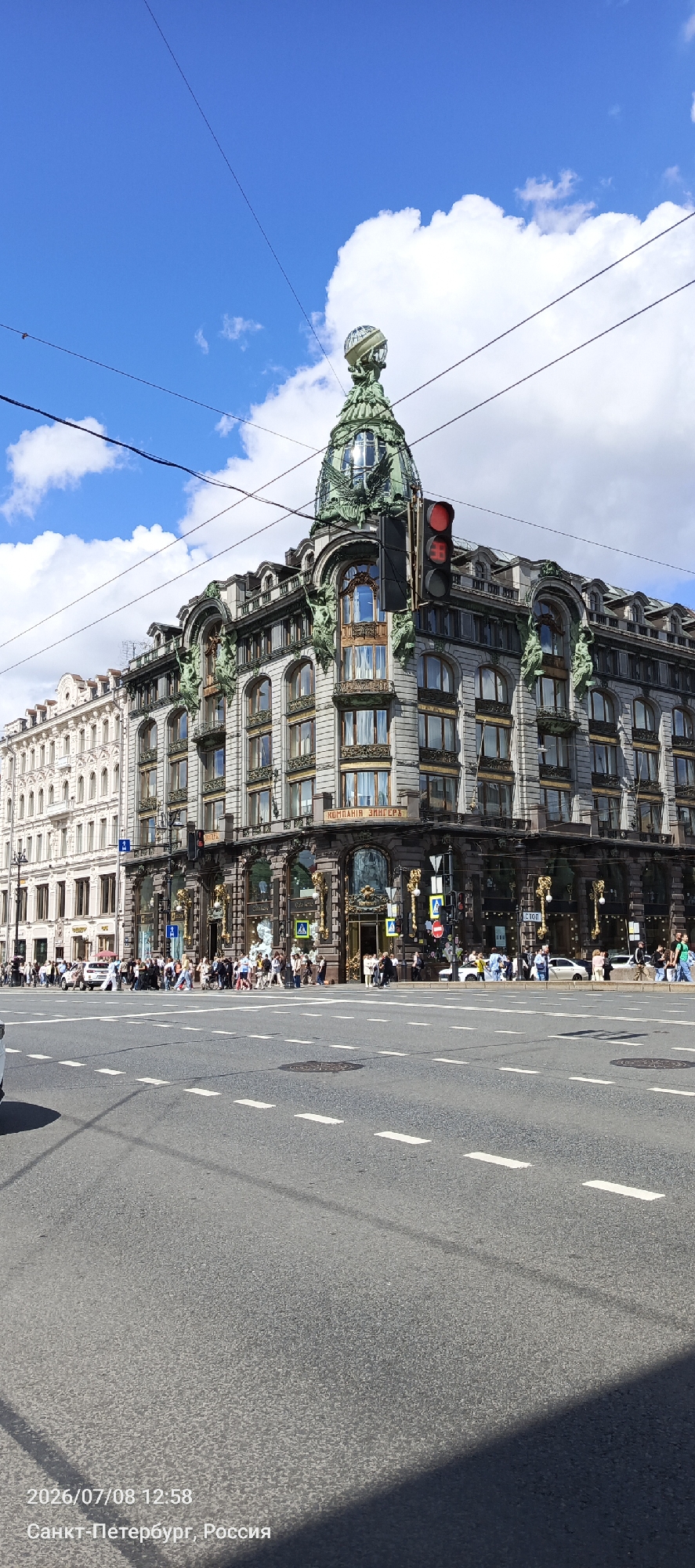 The image shows the Singer House (Dom Knigi) in Saint Petersburg, Russia, on a bright, partly cloudy day. The iconic building, known for its ornate architecture and glass globe on top, occupies the right side of the frame. It features multiple stories with arched windows, decorative sculptures, and intricate stonework. A prominent traffic light with a red signal is visible on the building's facade.
In front of the Singer House, a street with pedestrian crossings and road markings is present. A group of pedestrians is visible on the sidewalk and crossing the street, indicating a public area with moderate foot traffic. Several cars are also seen on the road. To the left of the Singer House, another building with a more classical architectural style is partially visible, presenting a contrast in design. The sky above is blue with scattered white clouds. The lower portion of the image shows the asphalt road surface, with white dashed lines marking lanes. The timestamp and location "2026/07/08 12:58 Санкт-Петербург, Россия" are overlaid in the bottom left corner of the image, indicating the date, time, and city of Saint Petersburg, Russia.