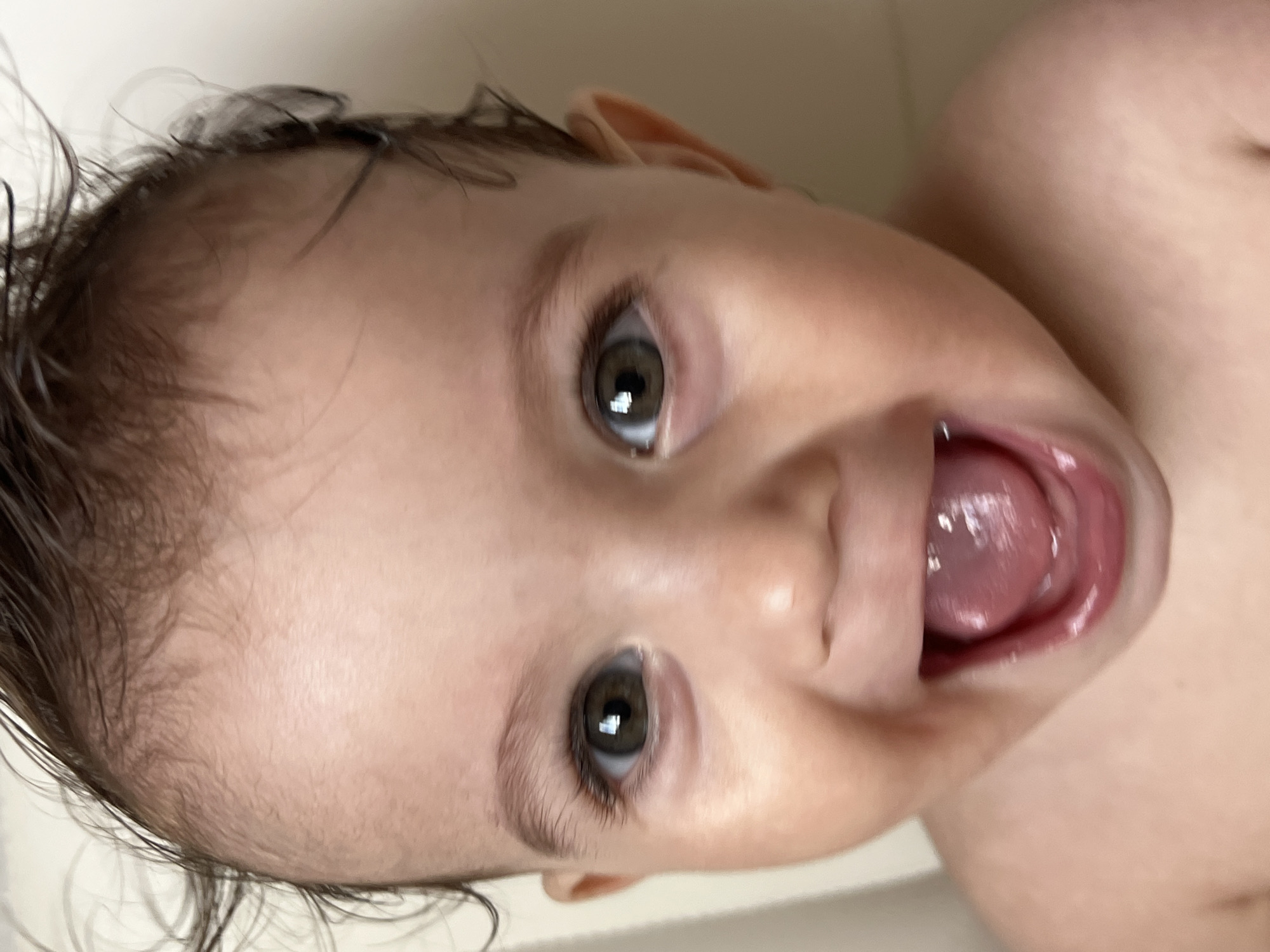 A close-up, off-center image shows a baby lying on its back. The baby's face is predominantly in focus, exhibiting dark hair and light skin. The baby's mouth is open. The background is blurry and indistinct. No information is visible regarding the location of Elena, Bulgaria.