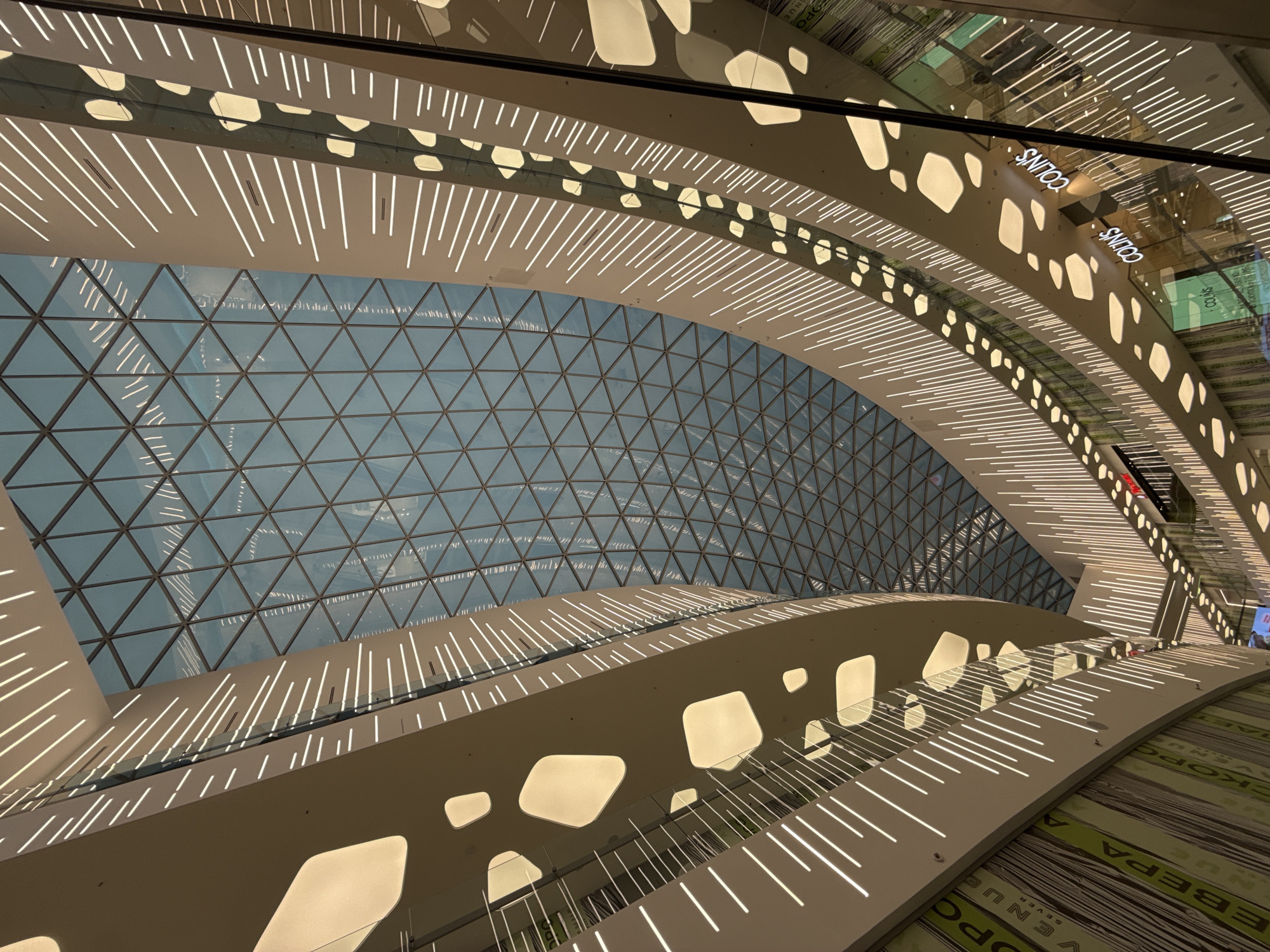 The image showcases the interior architecture of the Aviapark shopping mall in Moscow, Russia. The view is directed upwards, revealing a triangular grid-patterned glass ceiling, allowing natural light to filter through. The internal structure is comprised of multi-level curved balconies or walkways with integrated lighting. These walkways feature linear lighting elements along their edges and organically shaped cutouts that also emit light. At least one storefront is visible with the word "COLIN'S" displayed. The general atmosphere is brightly lit and modern.