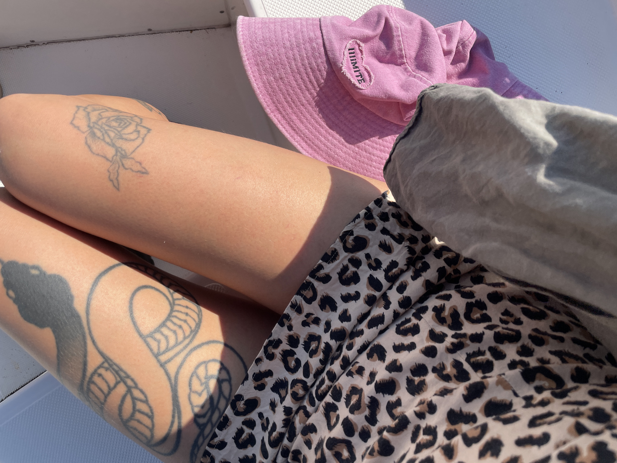 A close-up shot shows a person's legs adorned with tattoos. One leg has a large tattoo of a coiled snake, while the other features a delicate rose tattoo. The person is wearing a leopard-print skirt and a pink bucket hat with the word "LIMITE" embroidered on it. The scene is set outdoors, bathed in sunlight, suggesting a warm day. The background is a light-colored, textured surface, possibly a boat deck or outdoor seating. The overall impression is one of leisure and relaxation on a sunny day in Budva, Montenegro.