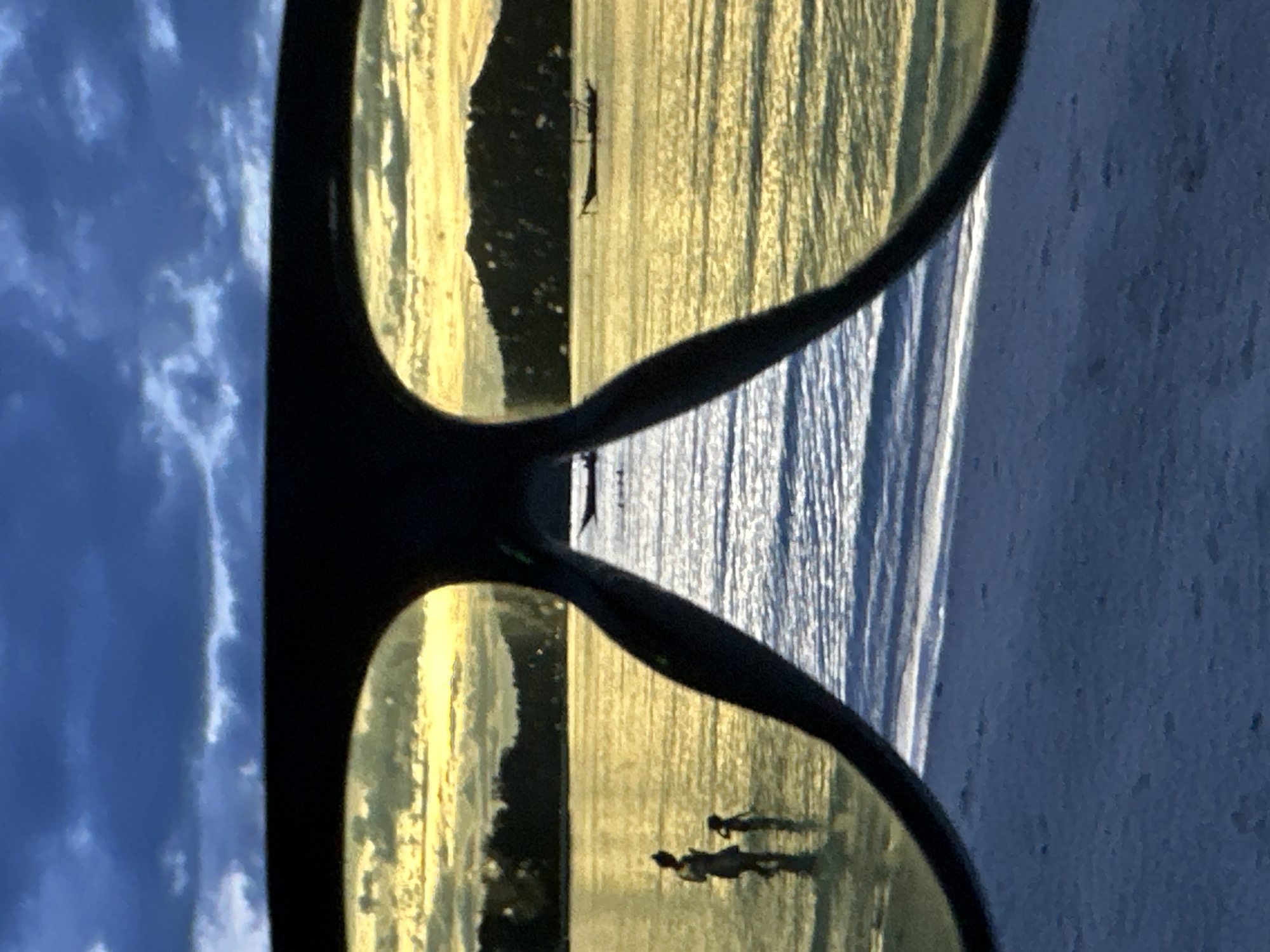 A pair of sunglasses reflects a coastal scene in ตำบลเกาะพะงัน, Thailand. The sunglass lenses show a beach at sunset, with figures of people walking on the shore. The ocean is visible in the reflection, with sunlight shimmering on the water. Outside the sunglasses, there is a view of the sky with clouds. The image is rotated.