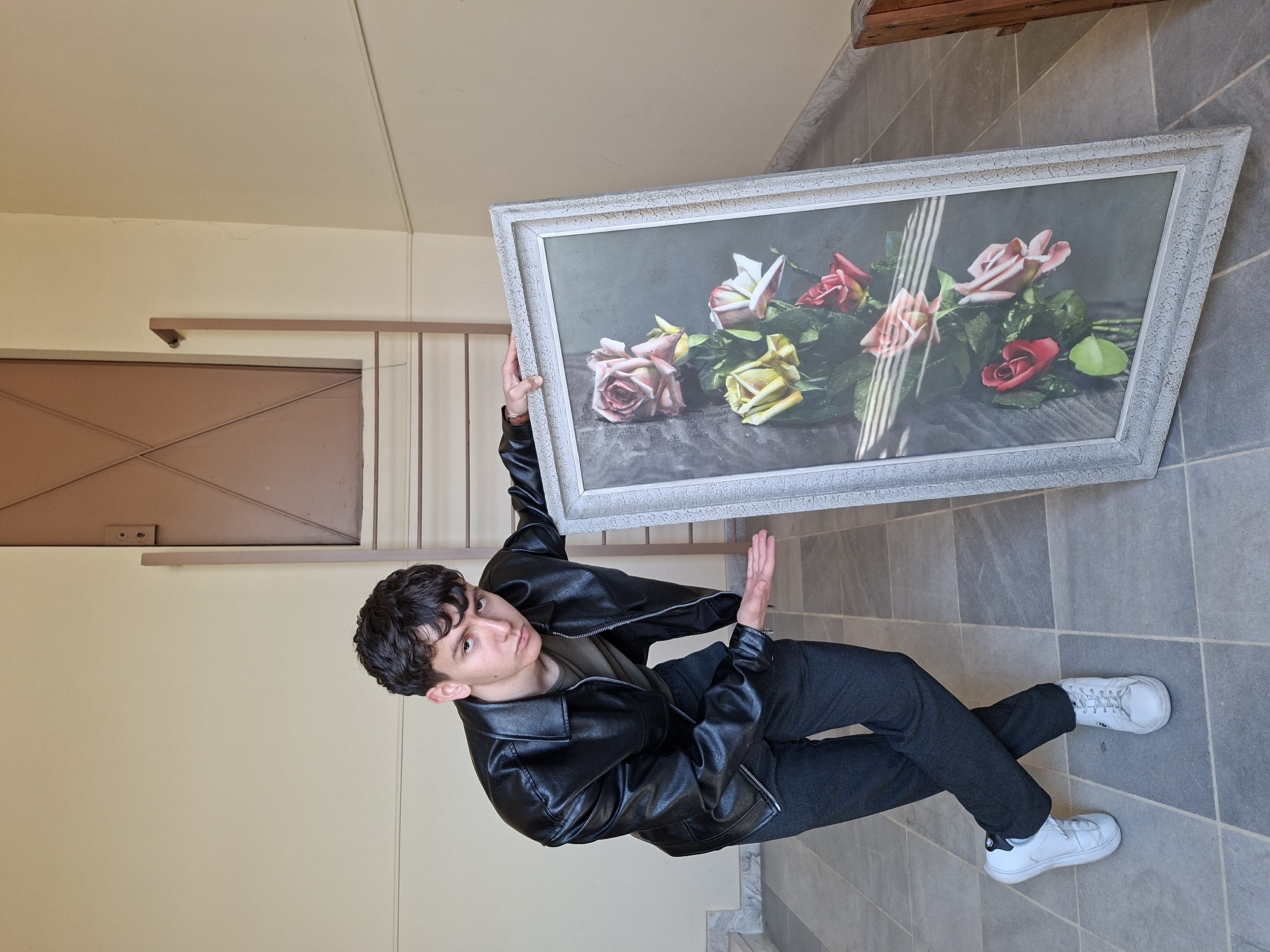 The media file features a young man, likely in his late teens or early twenties, standing indoors in what appears to be a hallway or stairwell. He has short, dark, curly hair and is dressed in a black faux-leather jacket over a dark olive-green t-shirt, dark gray pants, and white sneakers.
The main activity is the man holding a large, rectangular framed picture. The picture inside the distressed white or light gray frame displays a still life of several roses in shades of pink, yellow, and red, arranged on a dark, textured surface. Distinct vertical streaks of light, possibly from a window, are visible across the painting. The man supports the frame with his left hand behind it, while his right hand is extended, resting on the side of the frame or gesturing. He looks directly at the viewer with a neutral expression.
Notable details include the setting, which features a floor covered in gray square tiles, plain light beige walls, and a matching ceiling. In the background, a brown paneled door with an X-pattern and a simple railing are partially visible. The lighting suggests it is daytime. The image is taken indoors, in what is likely a building in Plovdiv, Bulgaria. There is no clearly legible text visible in the image.