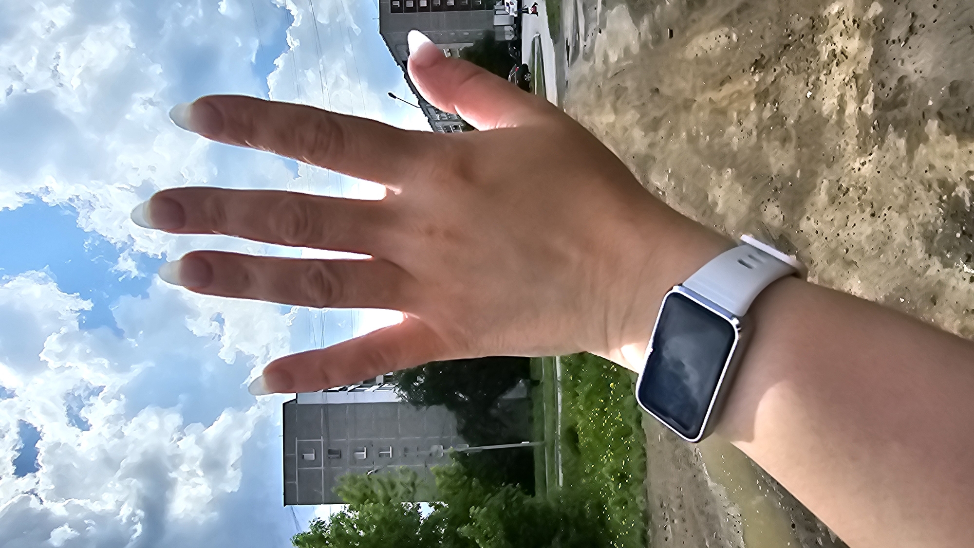 The image depicts a right hand, with long nails, extending towards the top left corner, a wrist adorned with a silver square smartwatch with a light grey band, and a scene suggesting an urban environment. The sky, filled with white clouds, appears in the top left corner behind the fingers. In the background are trees, an apartment building, a muddy patch of ground, and a street. The location is an unspecified area within a city. The focus is on the hand and wrist.