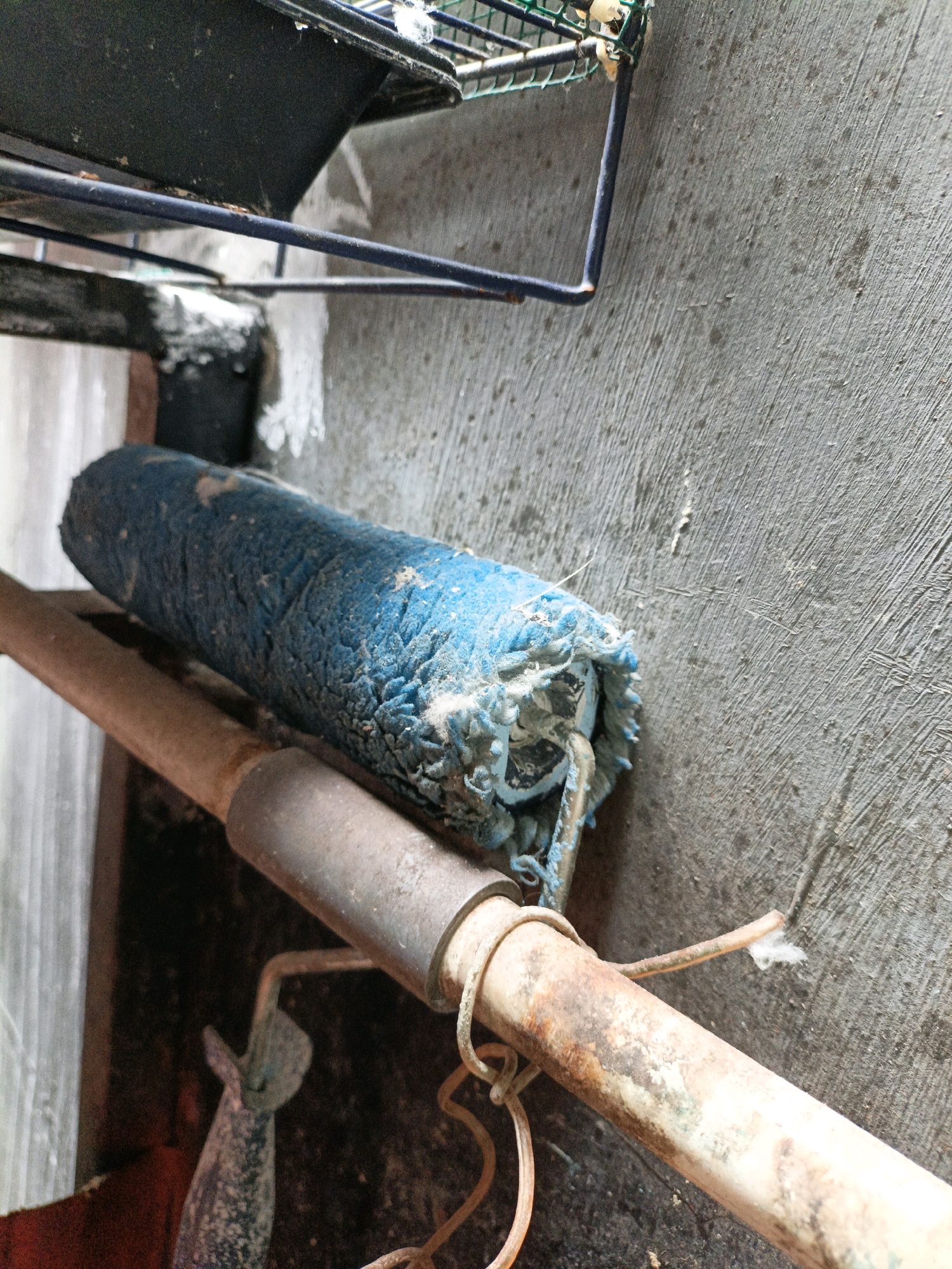 A close-up, low-angle shot shows a heavily used, blue paint roller lying horizontally on a rusty metal pipe. The roller is covered in dried paint residue and has frayed edges. The pipe itself is worn and discolored, with a thin wire wrapped around it. The background consists of a rough, textured concrete wall with visible brush strokes. A dark metal shelf or rack is visible at the top of the frame. The overall scene appears to be outdoors, possibly in a workshop or storage area. There are no people or active interactions depicted. The environment suggests a utilitarian setting in West Jakarta, Indonesia, where such tools are commonly found.