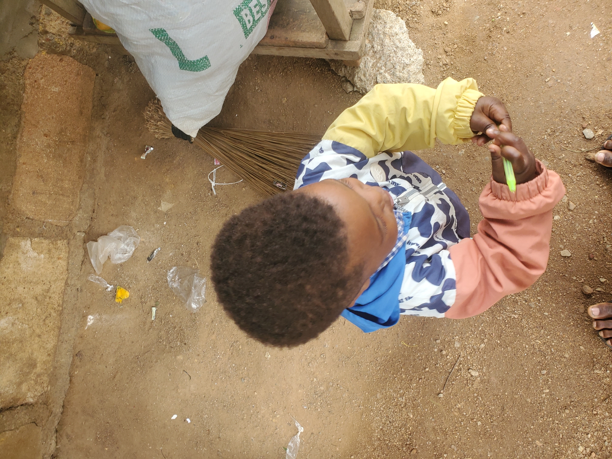 A young child, viewed from above, stands on sandy ground in what appears to be an outdoor area with a rough, stone or concrete wall in the background. The child is wearing a patterned jacket with blue, white, and dark blue camouflage print, a light blue hood, and long sleeves that are peach-colored on the left and bright yellow on the right. The child is holding a green object, possibly a pen or crayon, in their right hand. A broom made of dried grass is lying on the ground next to a large, white, woven sack with a green stripe. Several pieces of clear plastic debris and a yellow object are scattered on the ground. The ground is composed of fine sand and small pebbles. The overall impression is of a simple, possibly impoverished, outdoor setting. The question of whether this scene is located in Jos, Nigeria, is a speculative interpretation and not directly ascertainable from the visual information presented.