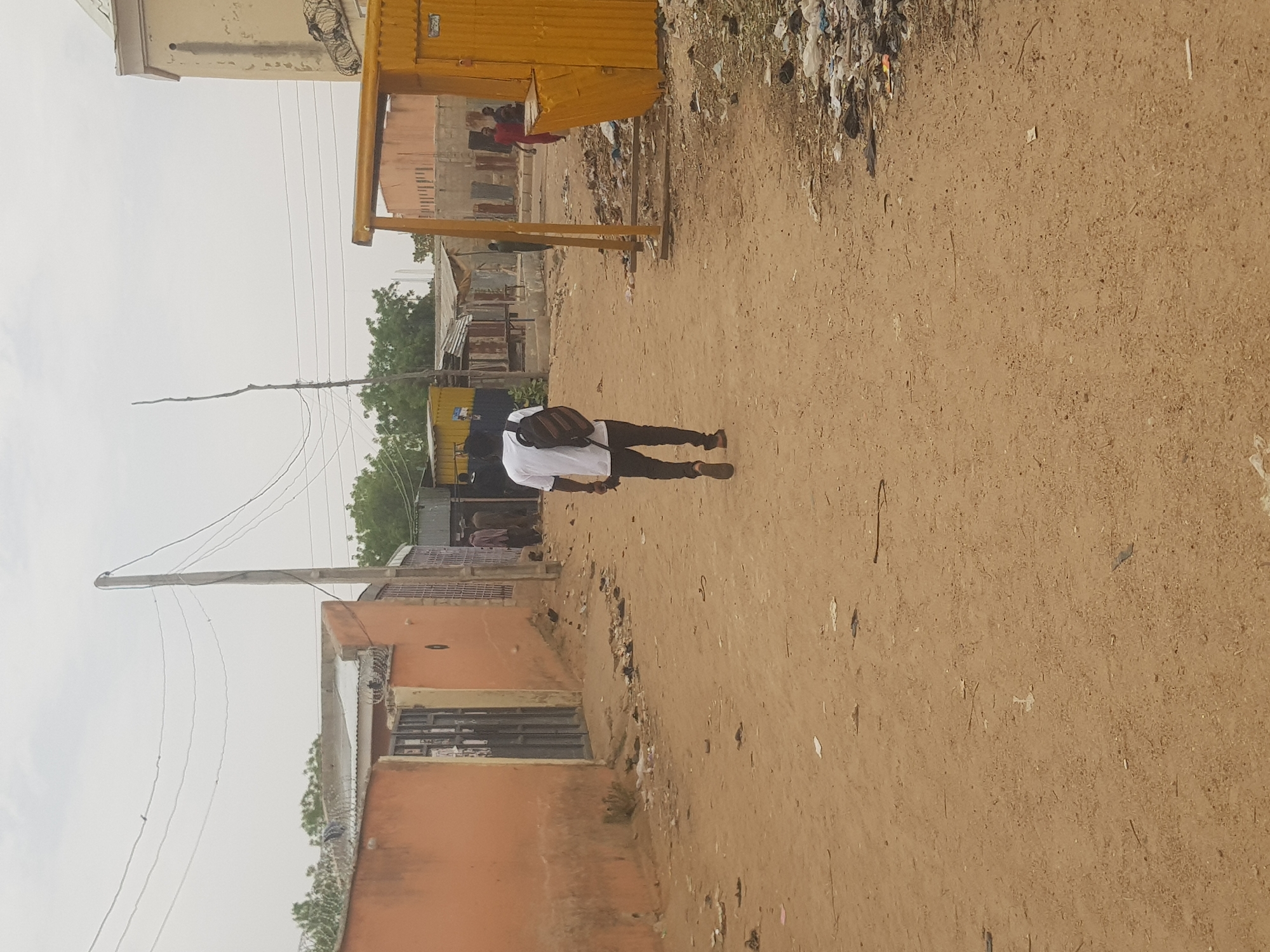 The image depicts an outdoor scene in Dabawa, Nigeria. A person wearing a white shirt, dark pants, and carrying a dark bag is walking away from the camera on a dirt road. To the right of the road is an area with litter. On the left are buildings, some made of orange material and others more makeshift. A yellow structure is also visible in the background. Overhead are power lines. The sky is overcast.