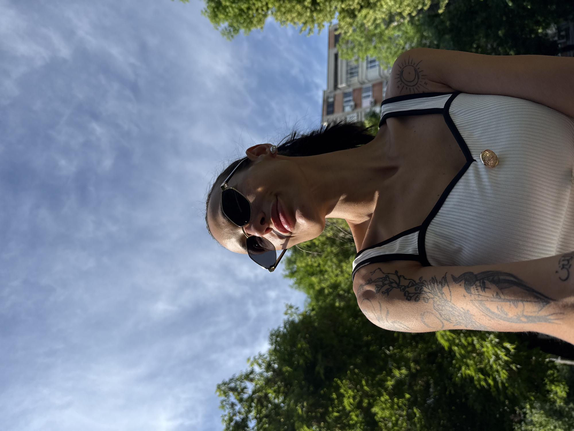 A woman wearing sunglasses and a white top with black trim stands outdoors. She has tattoos on both arms. In the background is a partly cloudy sky, trees, and a building. The location is Pleven, Bulgaria, determined based on the analysis of the environment and objects in the image.
