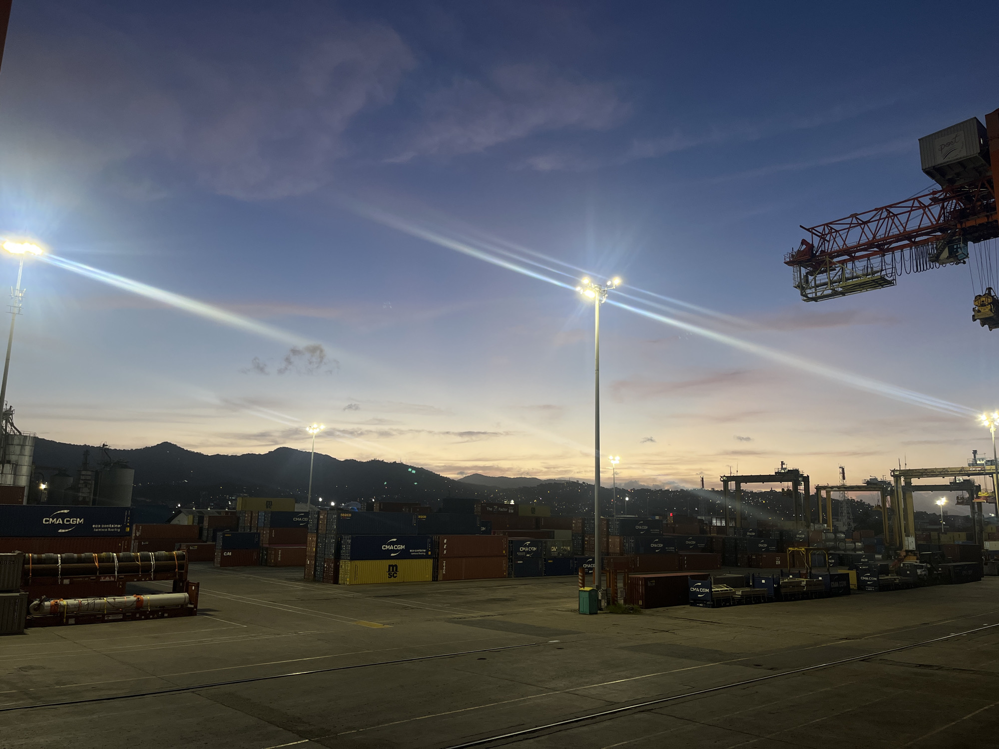 Here is a detailed description of the image:
**Scene:** The image depicts a port or shipping yard at twilight, likely located in a coastal city given the backdrop of hills or mountains in the distance. The sky transitions from a deep blue to a pale orange near the horizon.
**Objects:**
* **Shipping Containers:** Numerous shipping containers are stacked in rows across the scene. Many are labeled with shipping company logos and markings.
* **Cranes:** Large cranes, likely used for moving containers, are visible in the background on the right side of the image.
* **Light Poles:** Bright light poles illuminate the area with intense beams.
* **Pavement:** The foreground is dominated by a paved area, likely the operational surface of the port.
**Environment:**
* **Lighting:** The ambient light is low, with the sky in twilight and the port area lit by artificial lights.
* **Atmosphere:** The atmosphere seems clear.
* **Landscape:** Hilly or mountainous terrain forms the backdrop of the scene.
**No people are discernible in the image.**