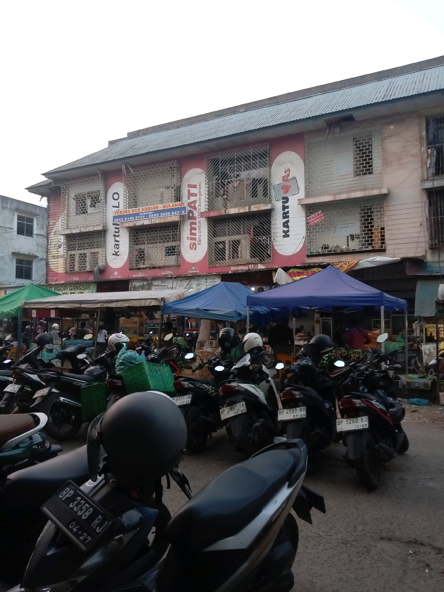 A street scene in Batam, Indonesia, features a row of parked motorcycles, predominantly scooters, in the foreground and midground. Behind the motorcycles, market stalls covered by blue and green awnings are visible, with people moving around within the market area. A multi-story building, painted pink and white, forms the backdrop. The building's facade has several windows with metal grates and prominent advertisements with text in Indonesian, including "KARTULO" and "simPATI." A sign with the text "TERIMA KOS HARIAN - BULANAN" is also visible. The sky above is overcast and a light grey color. The ground is paved with asphalt, showing signs of wear.