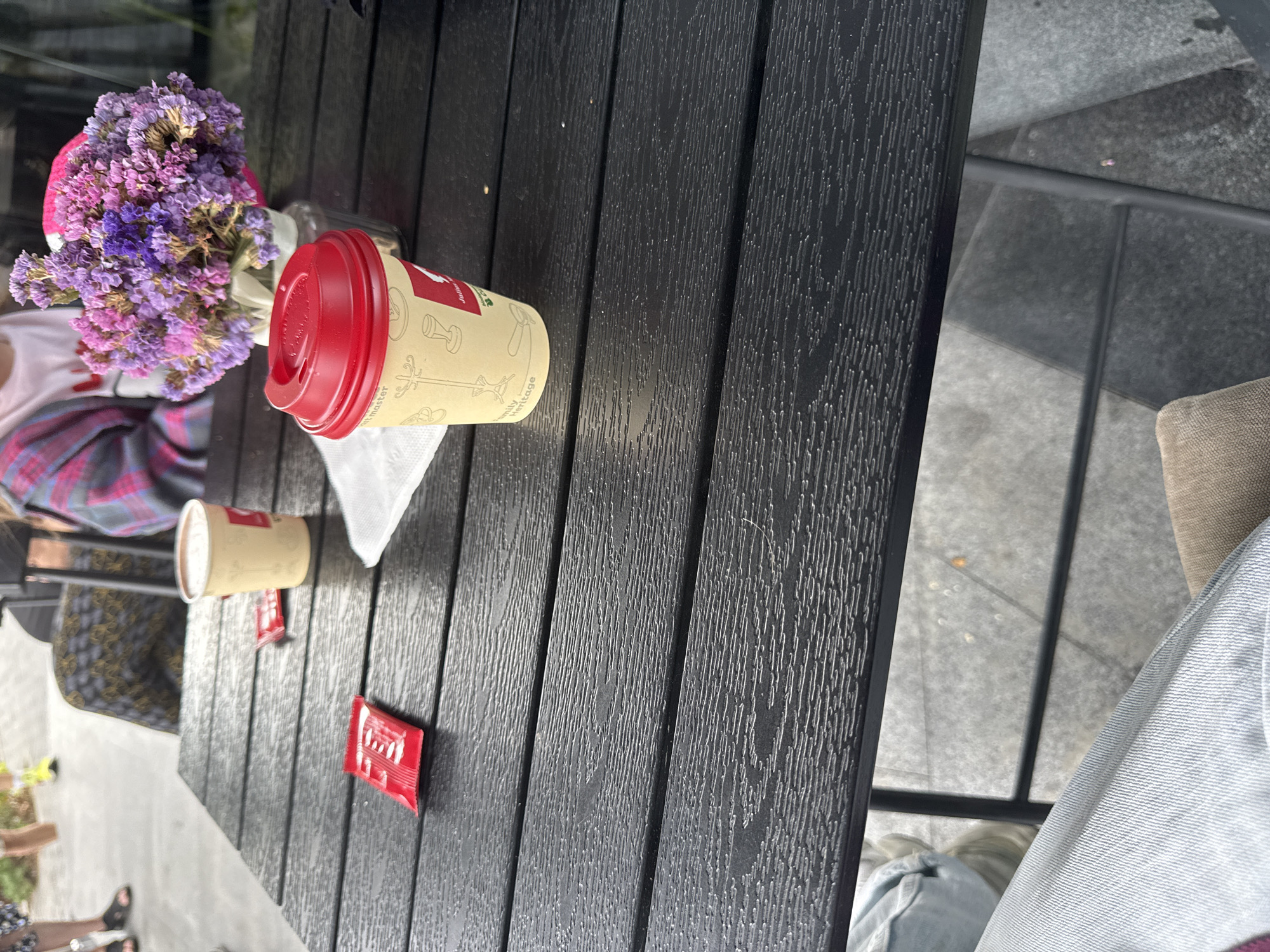 A high-angle outdoor shot shows a black wooden table with two disposable coffee cups and a small bouquet of dried flowers in a vase. One cup, topped with a red lid, is closer to the foreground. A red packet, likely containing sugar or creamer, lies to the left of this cup. Further back on the table, another, partially obscured disposable cup is visible, along with another red packet. The table is set on an outdoor patio area with a grey paved ground. In the background, blurred figures of people and a patterned chair can be seen. The overall scene suggests a relaxed outdoor cafe setting, potentially in Chișinău, Moldova, given the user's prompt.