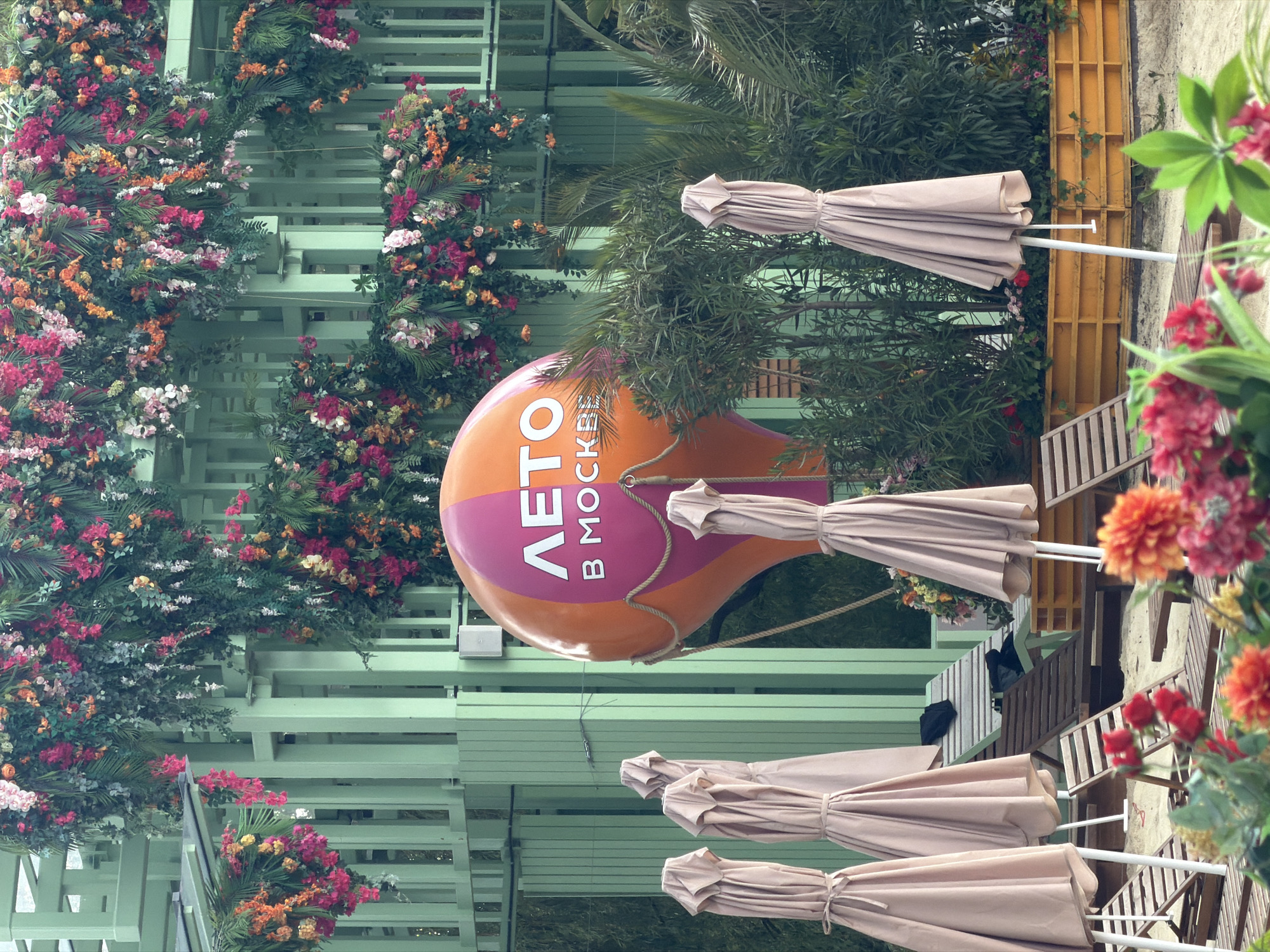 The image captures an outdoor space in Moscow, Russia, decorated for a summer theme. A sign shaped like a hot air balloon is visible, with the Russian words "ЛЕТО В МОСКВЕ," which translates to "Summer in Moscow." The space appears to be a patio or outdoor dining area with tables and folded beige umbrellas. The architecture includes light green wooden structures adorned with vibrant floral arrangements. The scene suggests a casual and festive atmosphere, possibly a restaurant or event venue. The composition is a mix of natural and constructed elements, featuring foliage, flowers, and man-made structures. The image seems to have been taken during the day.