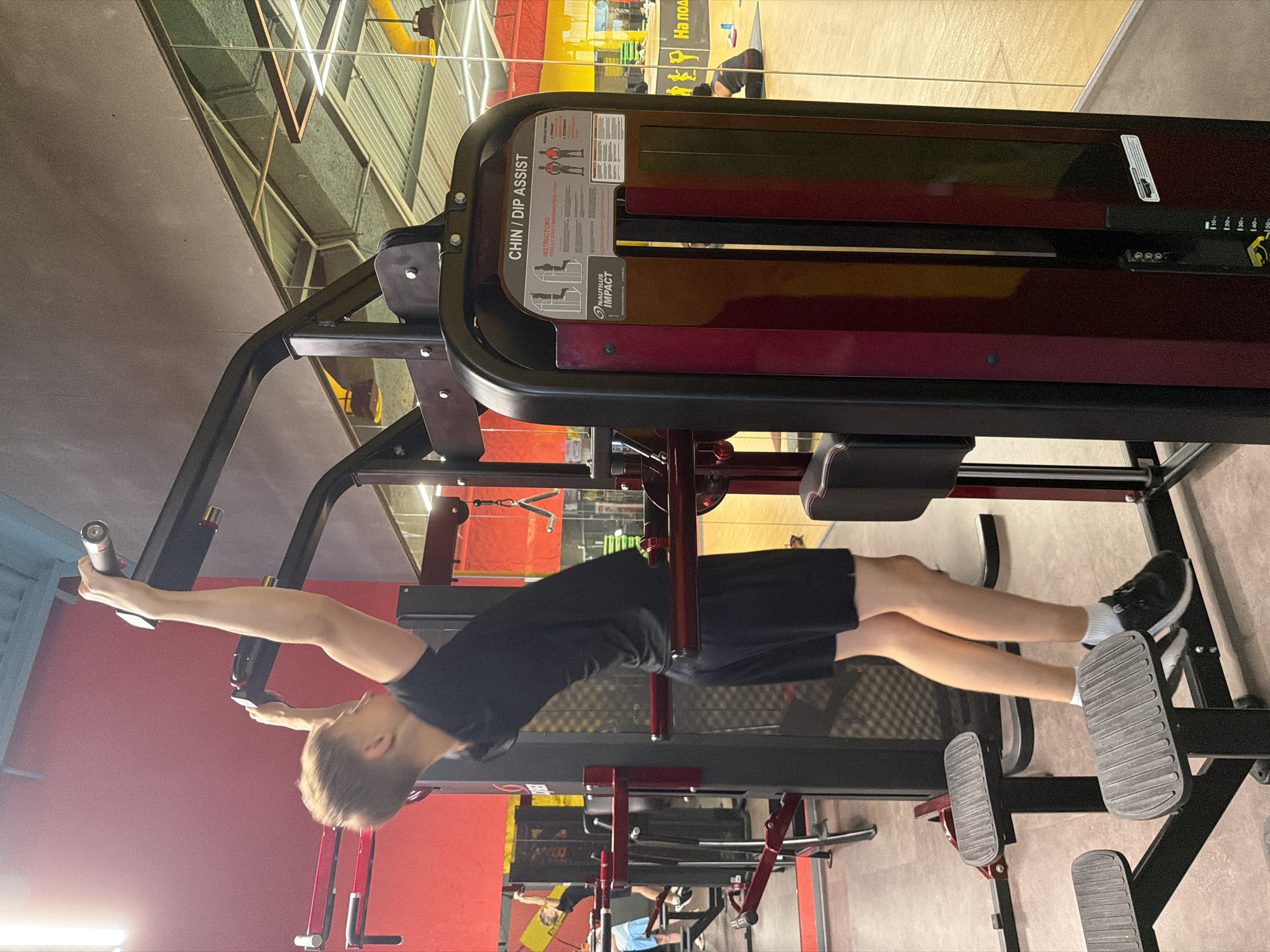 The image shows a young male performing a chin-up exercise on a Nautilus Impact Chin/Dip Assist machine in a gym. He is wearing a black t-shirt and black shorts. The machine is burgundy and black, with a weight stack visible on the side. The gym environment features red walls, mirrors, and other gym equipment in the background. The scene appears to be indoors. The time of day is not discernible. There are no discernible Kyiv-specific landmarks or indicators in the image.