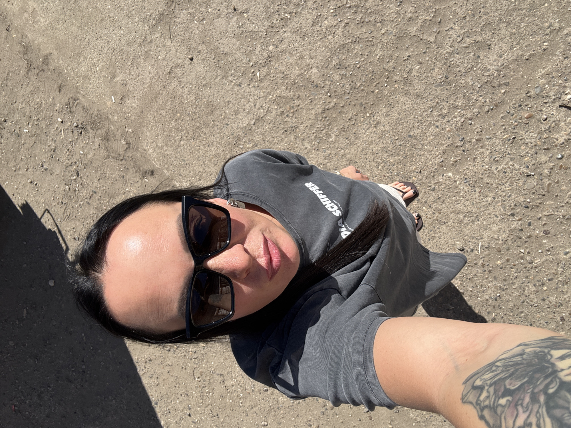 A high-angle selfie shows a woman standing on a dirt-covered surface. The woman has dark hair, sunglasses, and is wearing a grey t-shirt with "SCHIFFER" printed on it. Her right arm, visible in the foreground, has a tattoo. Her feet are clad in flip-flops. The dirt surface appears to contain pebbles and small debris. In the upper left corner is a shadow cast by an unknown object, possibly a building. This photo may have been taken in Chaikovsky, Russia.