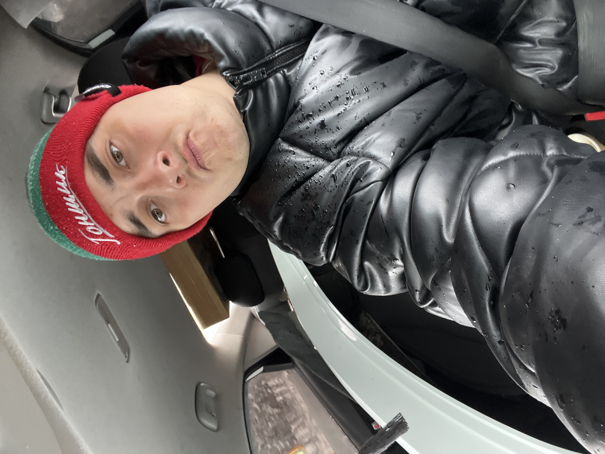 A man is captured in a close-up portrait, seemingly a selfie taken from a low angle, inside a car. He is wearing a red and green knitted beanie and a black puffer jacket that is noticeably wet with small water droplets, suggesting recent exposure to precipitation like rain or melting snow. A black seatbelt is visible across his chest.
The man looks directly at the camera with a slightly puckered expression. His eyes are brown. The red section of his beanie features white embroidered text, which clearly reads "Ширяево," confirming the location as Ширяево, Russia.
The car's interior is visible, including the light gray headliner above him with various recesses. To the right of the frame, a light-colored trim piece and a darker area, possibly the car's trunk opening or rear window, are seen, with faint indications of an outdoor, potentially wintery, scene. A cardboard-colored object, possibly a box, is partially visible behind his head on the left. The lighting is diffused, consistent with an overcast day.