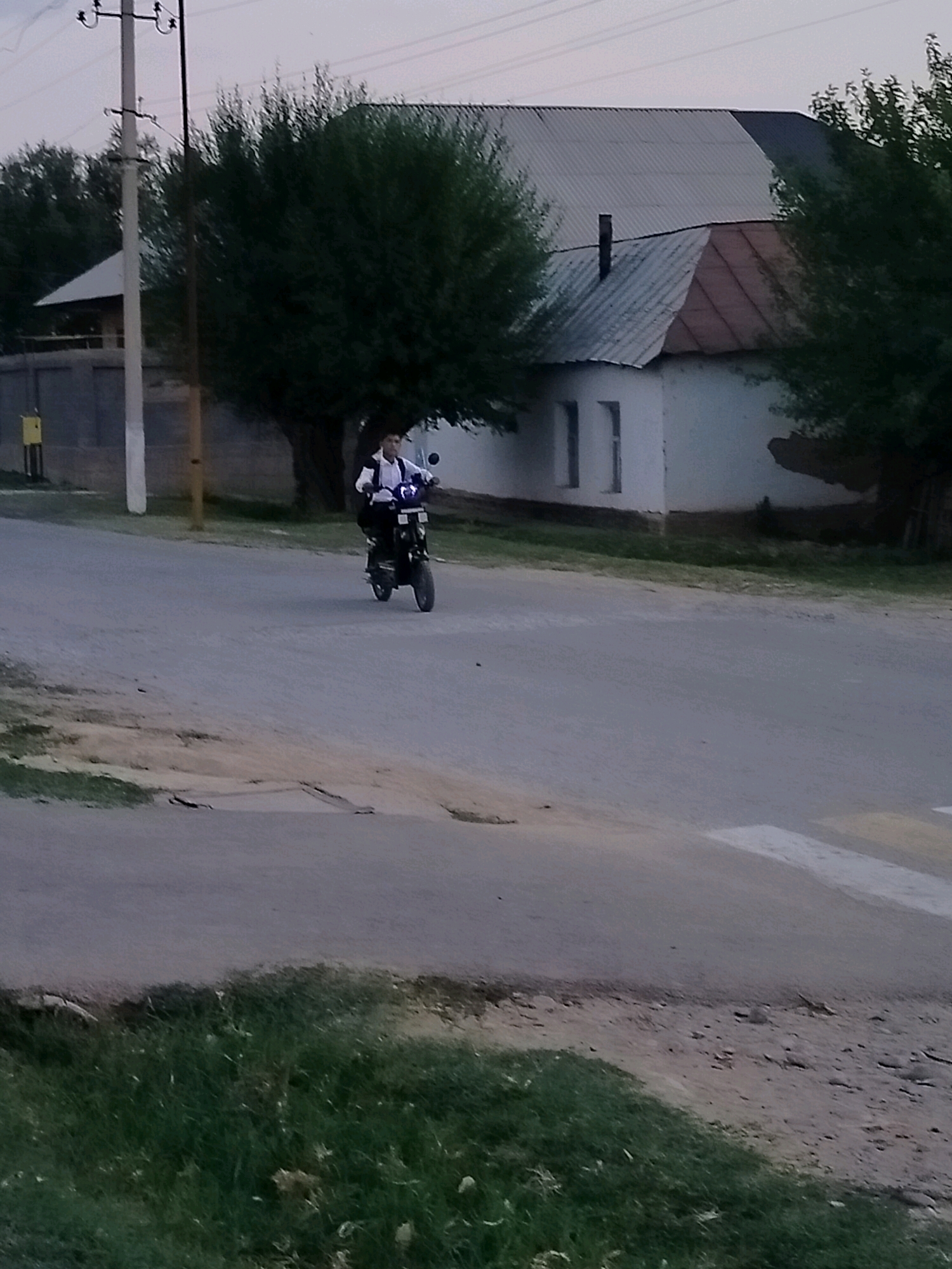 A young person wearing a white shirt and dark vest rides a dark-colored scooter on a paved road in a rural setting. The scooter has a headlight and mirrors. The person appears to be looking forward. The road is flanked by grass and dirt on the left and right sides. In the background, there is a white building with a corrugated metal roof, several trees, and a utility pole. The sky is overcast, suggesting it might be early morning or late evening, or perhaps a cloudy day. The overall scene depicts a calm moment in a residential area, possibly in Kazakhstan, given the location context. There are no visible text elements in the image.