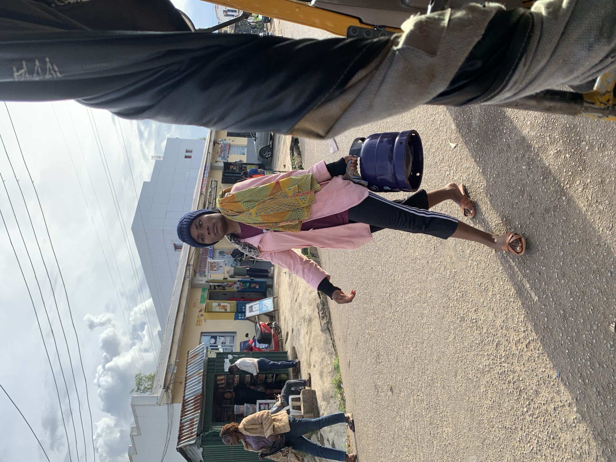 A woman wearing a blue beanie, a pink jacket, and dark pants with white stripes walks across a paved street. She carries a dark blue gas cylinder in her right hand and a patterned scarf is draped over her left shoulder. She is walking away from a row of shops with awnings and signs, one of which reads "POS". The street is lined with buildings and there are several people in front of the shops, some sitting and others standing. A red scooter is parked near one of the shops. In the foreground, a portion of a yellow vehicle, possibly a tuk-tuk or tricycle, is visible on the right side of the image. The sky is partly cloudy. The scene is likely set in the city of Jos, Nigeria, as indicated by the presence of local architecture and transportation.