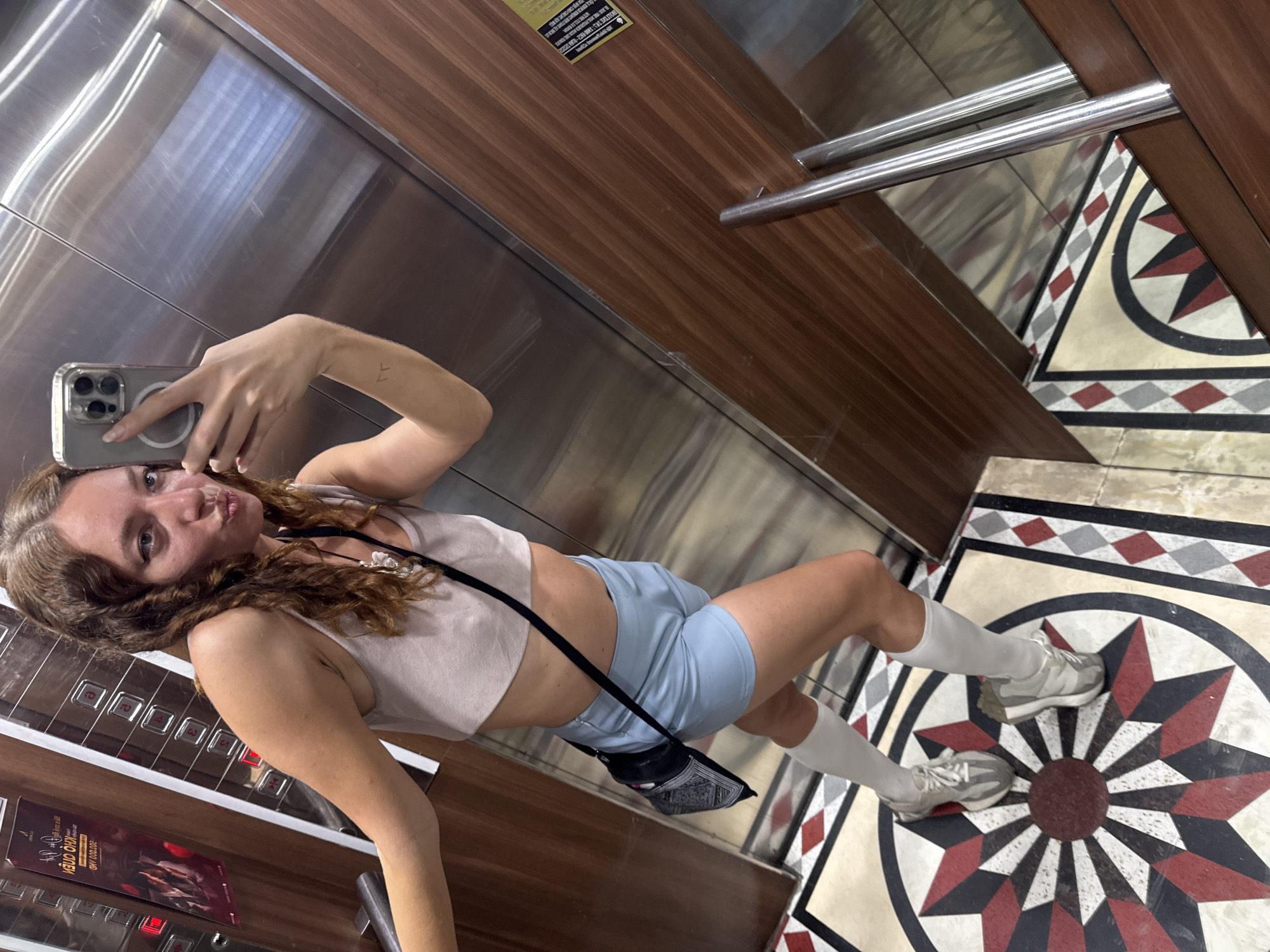 In an elevator with mirrored stainless steel walls and dark wood paneling, a young woman is taking a selfie. She is wearing a light pink crop top, light blue shorts, and white knee-high socks with grey sneakers. A small black bag with a patterned design hangs from her shoulder. Her right arm is extended, holding a silver iPhone, and she is puckering her lips towards the camera. A small tattoo is visible on her left forearm. The elevator floor has a decorative geometric pattern in shades of cream, red, black, and grey. A metal handrail runs along the wood-paneled wall. A poster with text in Vietnamese is visible on the elevator wall. The lighting suggests it is daytime. The overall mood appears casual and playful.